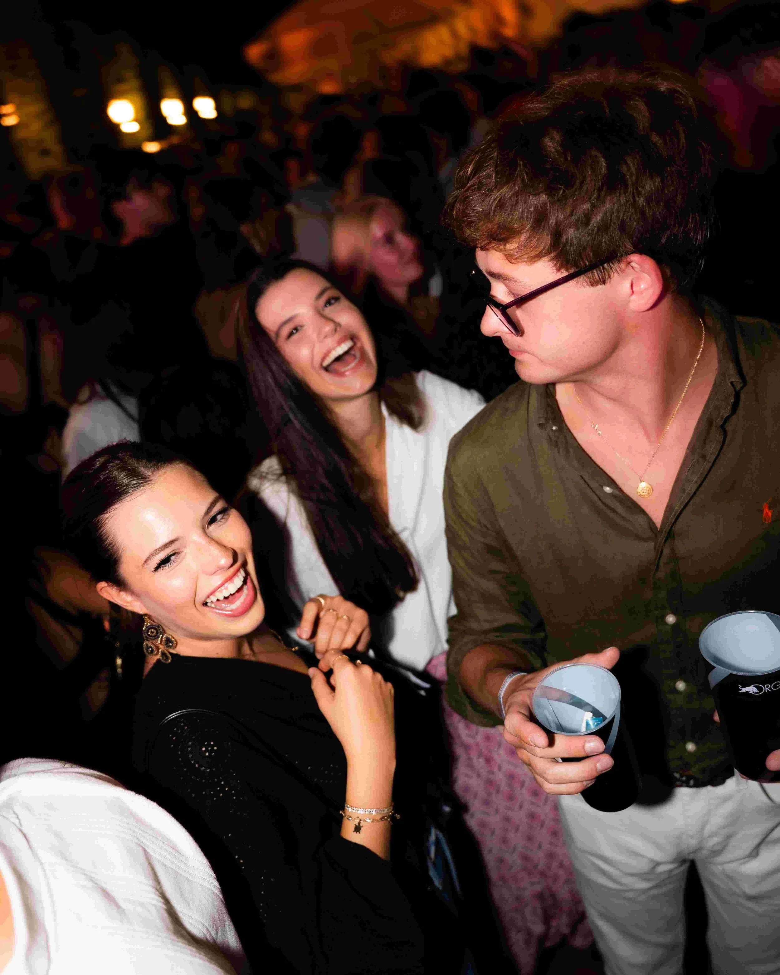 Group of friends at a party, smiling and having fun. One woman with dark hair and earrings sitting on a white couch, another woman laughing in the background, and a man with glasses holding drinks in his hands.