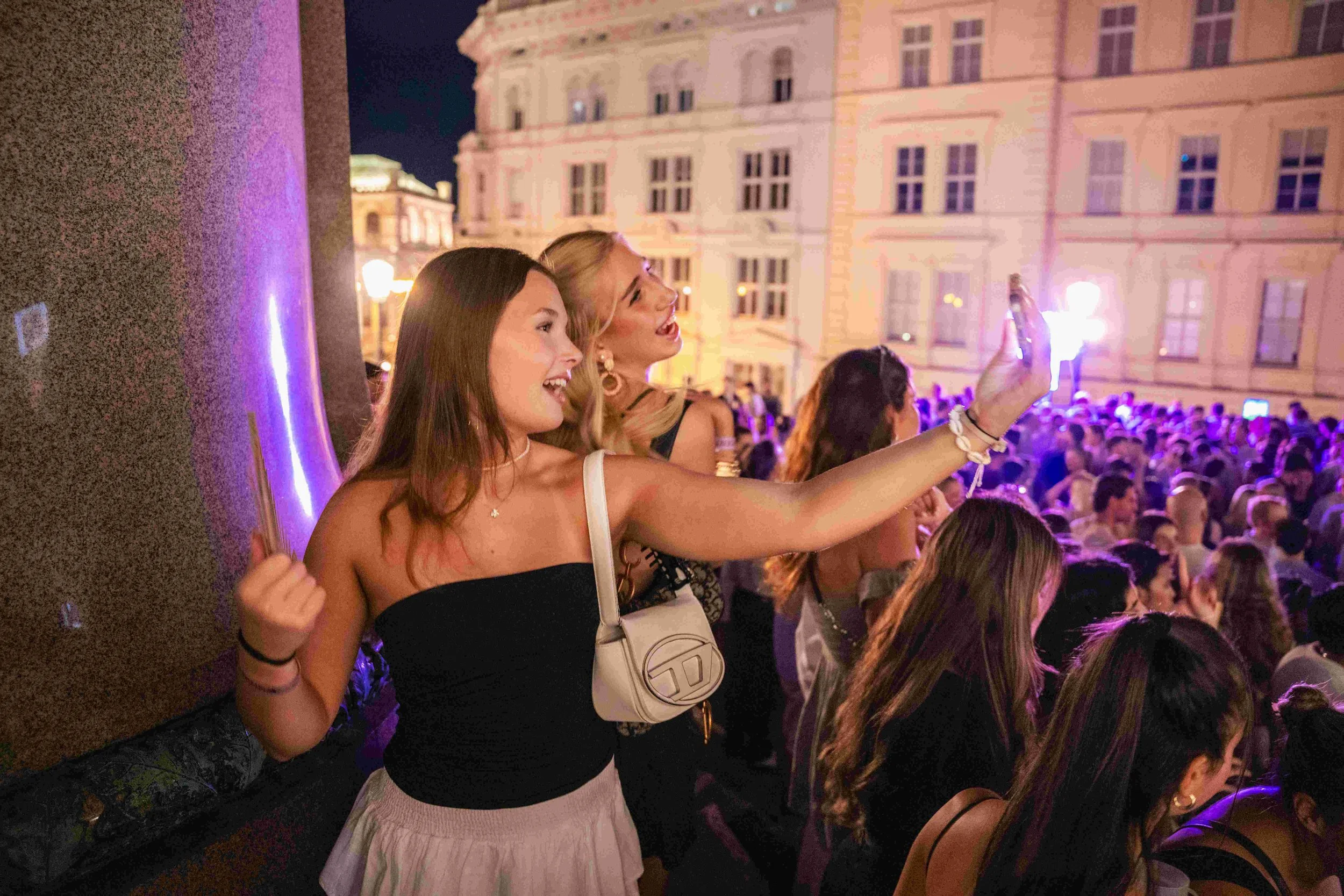 Two women smiling and taking a selfie at a crowded nighttime outdoor event in an urban setting.