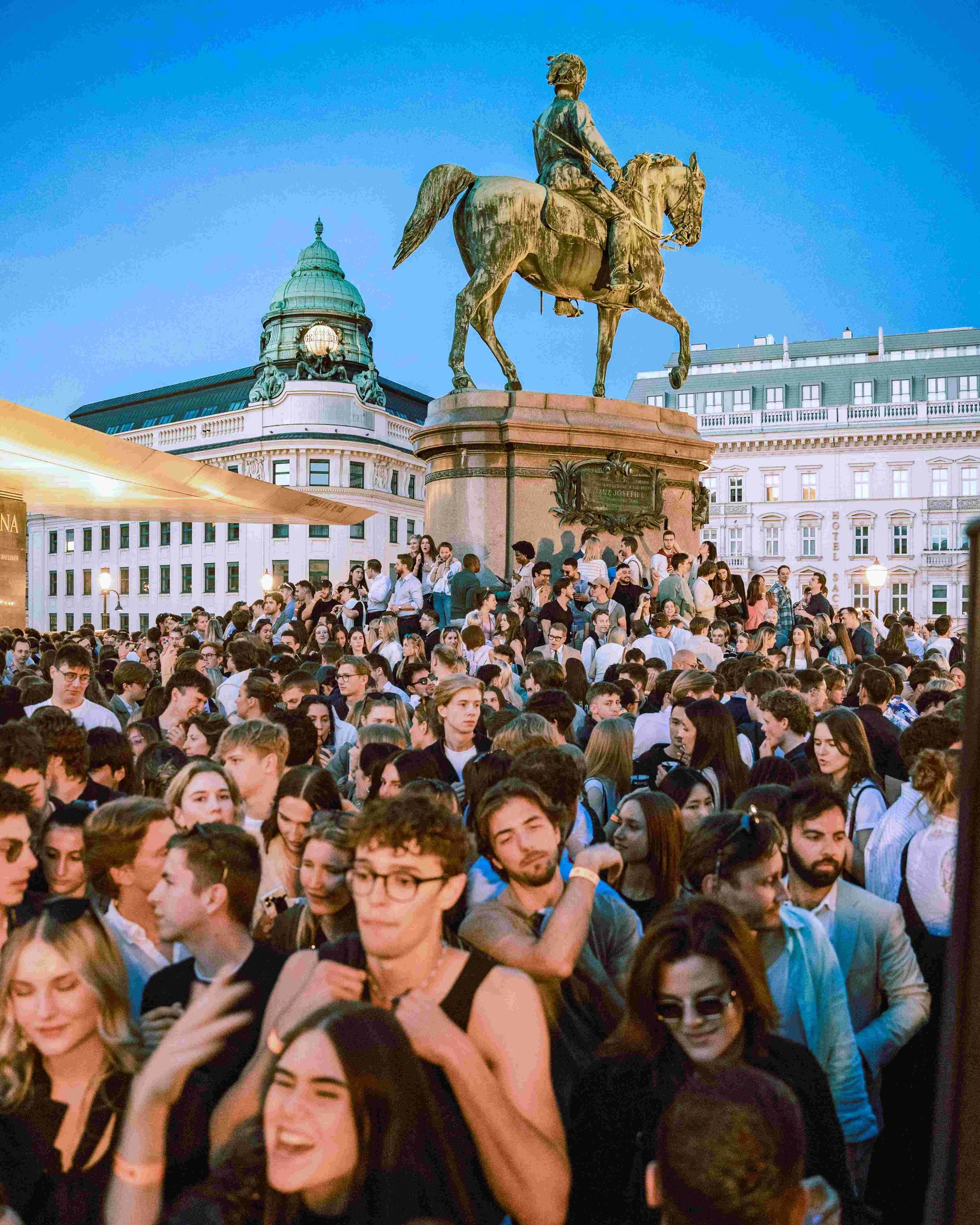 A large crowd of young people gathered outdoors in front of a historic building with a domed roof and a statue of a man on horseback. The sky is clear and blue, indicating it is either evening or early night.