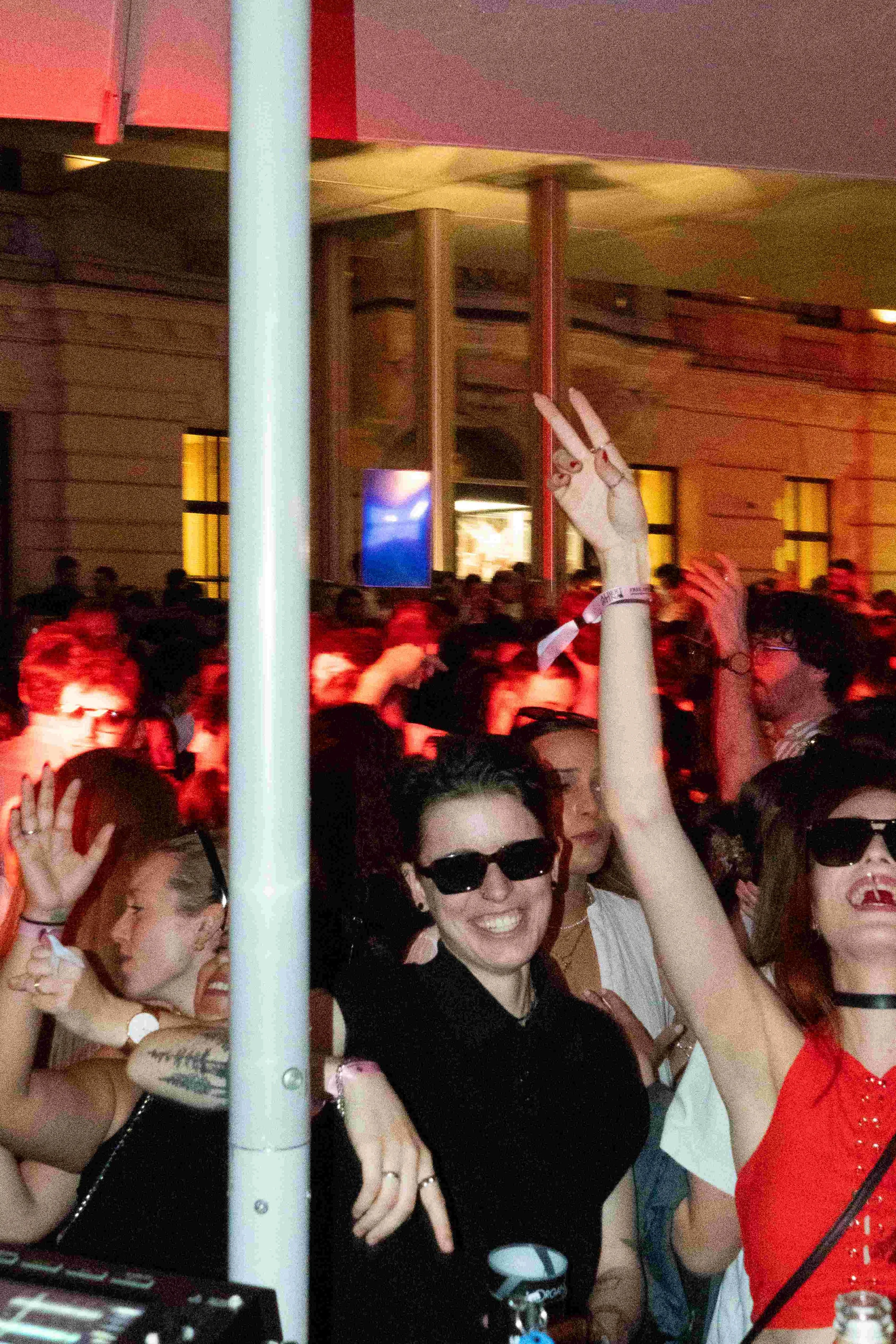 A crowd of people at a lively event or party. Some are wearing sunglasses, and one woman is smiling and making a peace sign. There is a person with a rabbit ear hat in the background. The setting appears to be indoors or under a tent with warm lighti