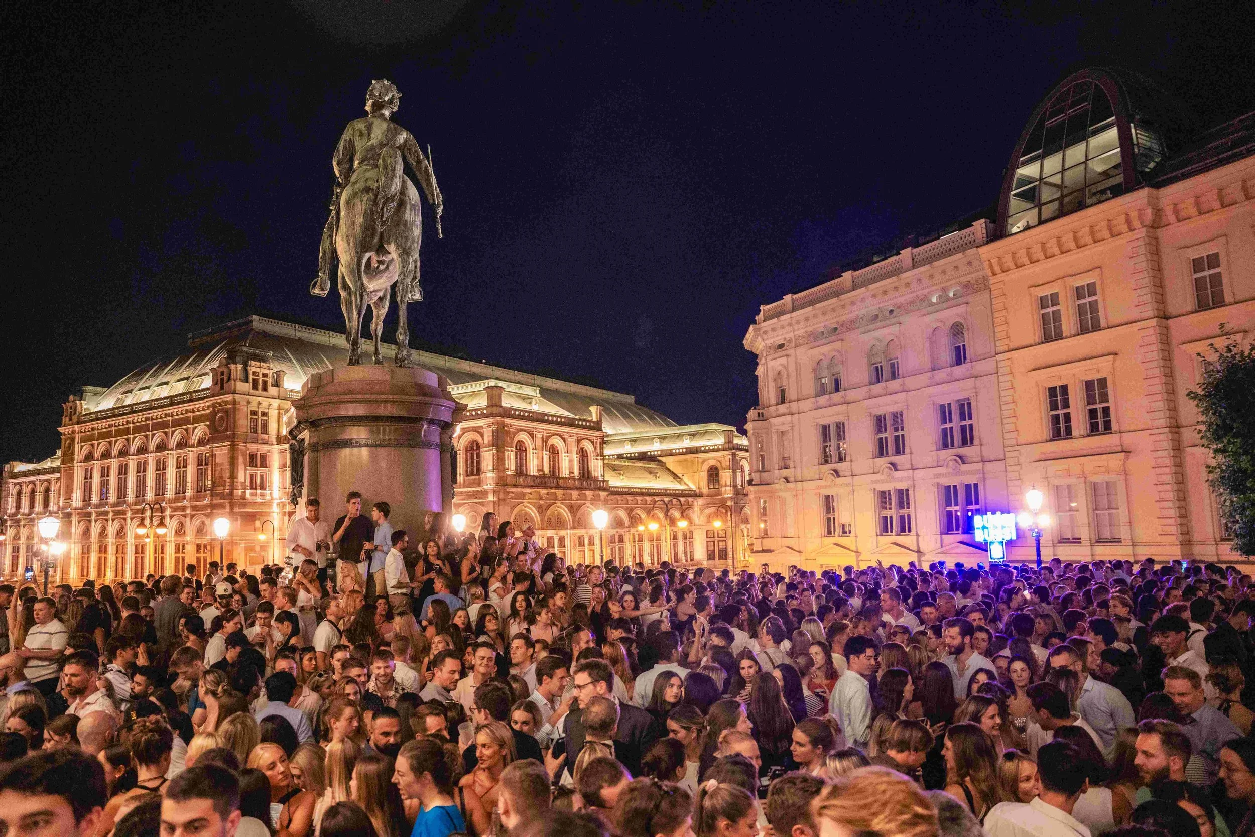 Nighttime scene of a crowded outdoor event with a statue of a man on a horse in the background, illuminated historic buildings, and many people gathered in the street.