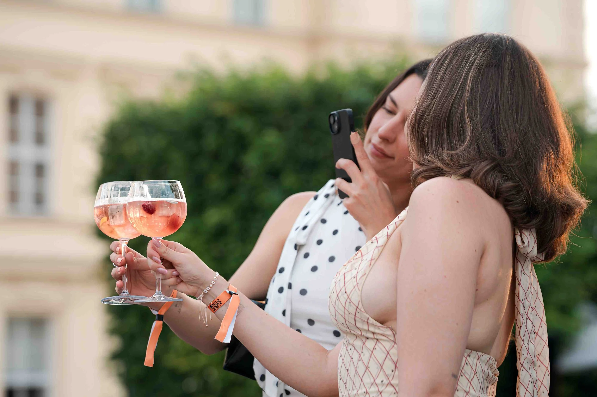 Two women at an outdoor gathering holding glasses of pink rosé wine with strawberries, one woman is taking a photo with her phone, both smiling and dressed in summer clothing.