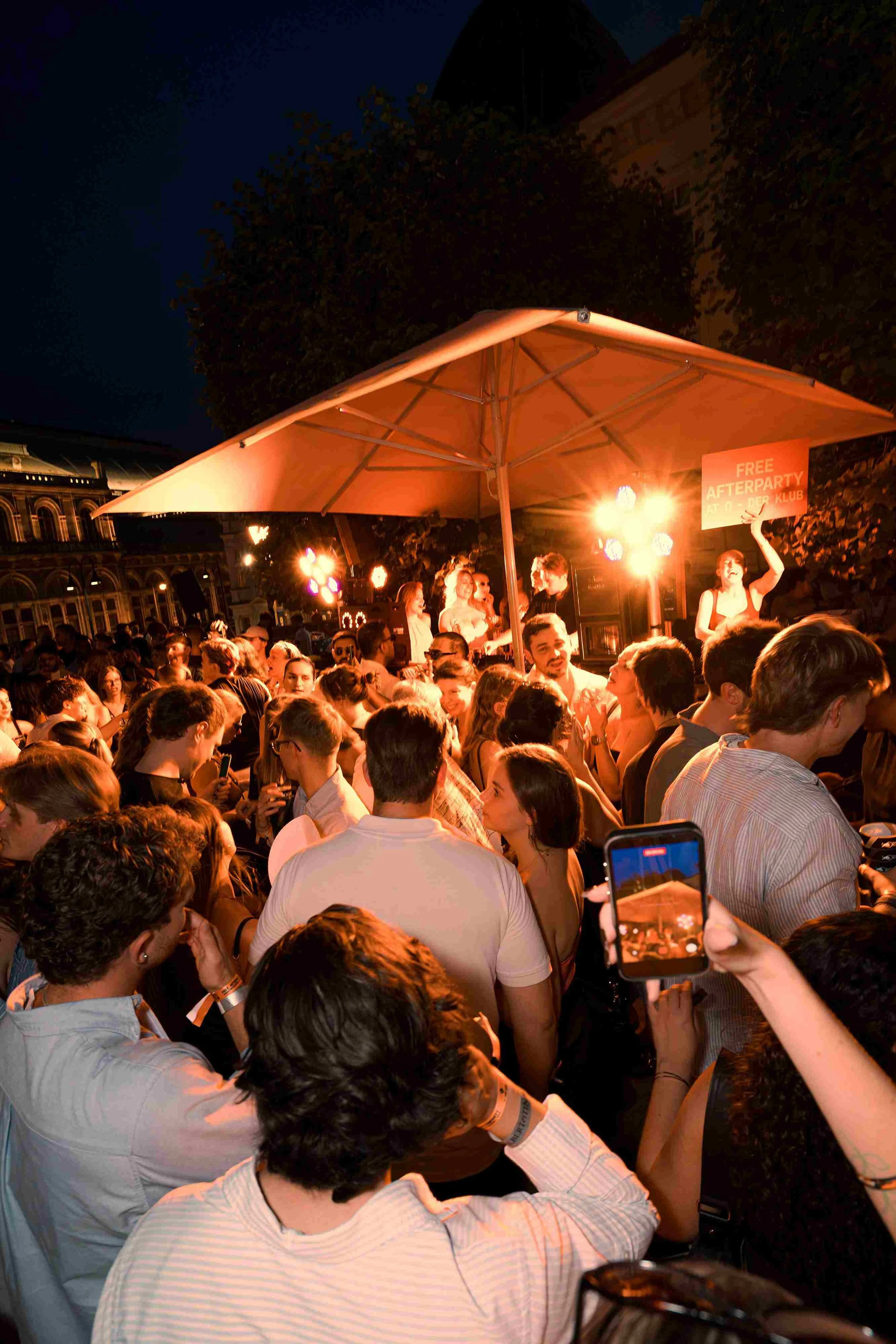 Crowd gathered at night outdoors with a woman holding a sign that reads 'FREE AFTERPARTY AT O DER KLUB,' under a large umbrella with lights behind them.