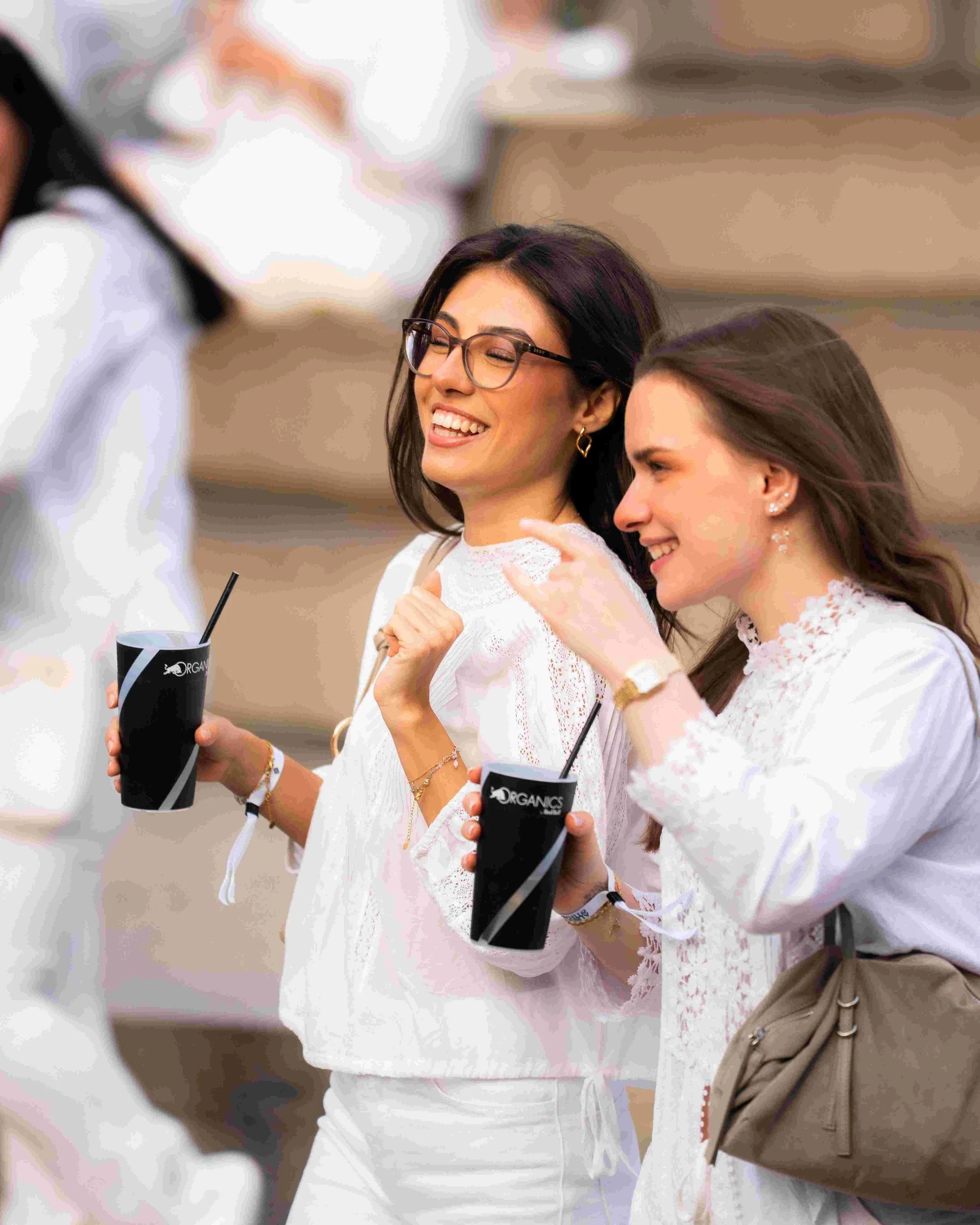 Two smiling women with brown hair and glasses, wearing white clothing, holding black cups with straws, outdoors with blurred background.