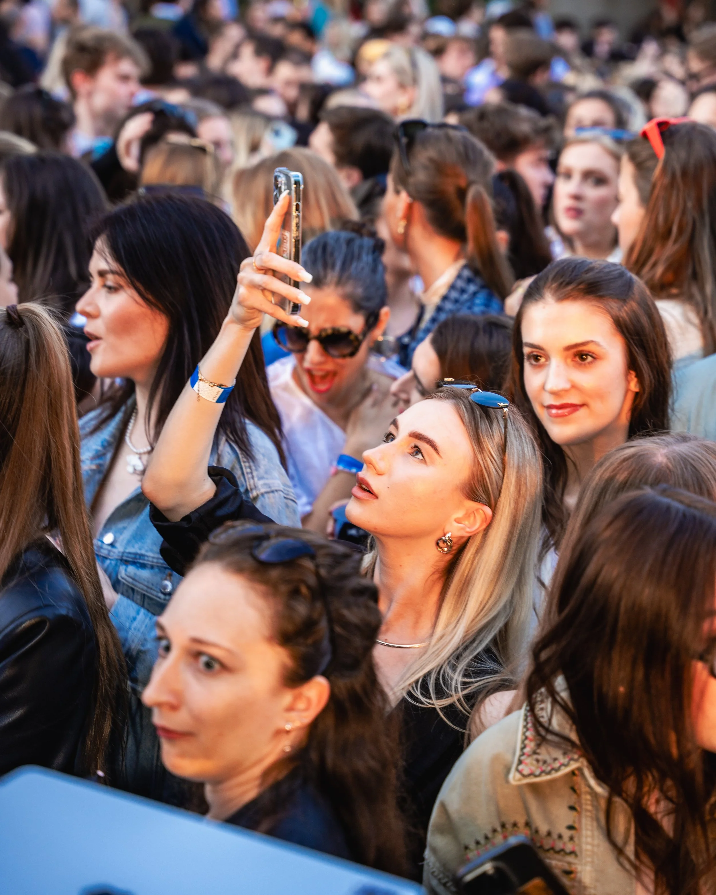 A large crowd of people gathered closely together, many of whom are taking photos or selfies with their smartphones.