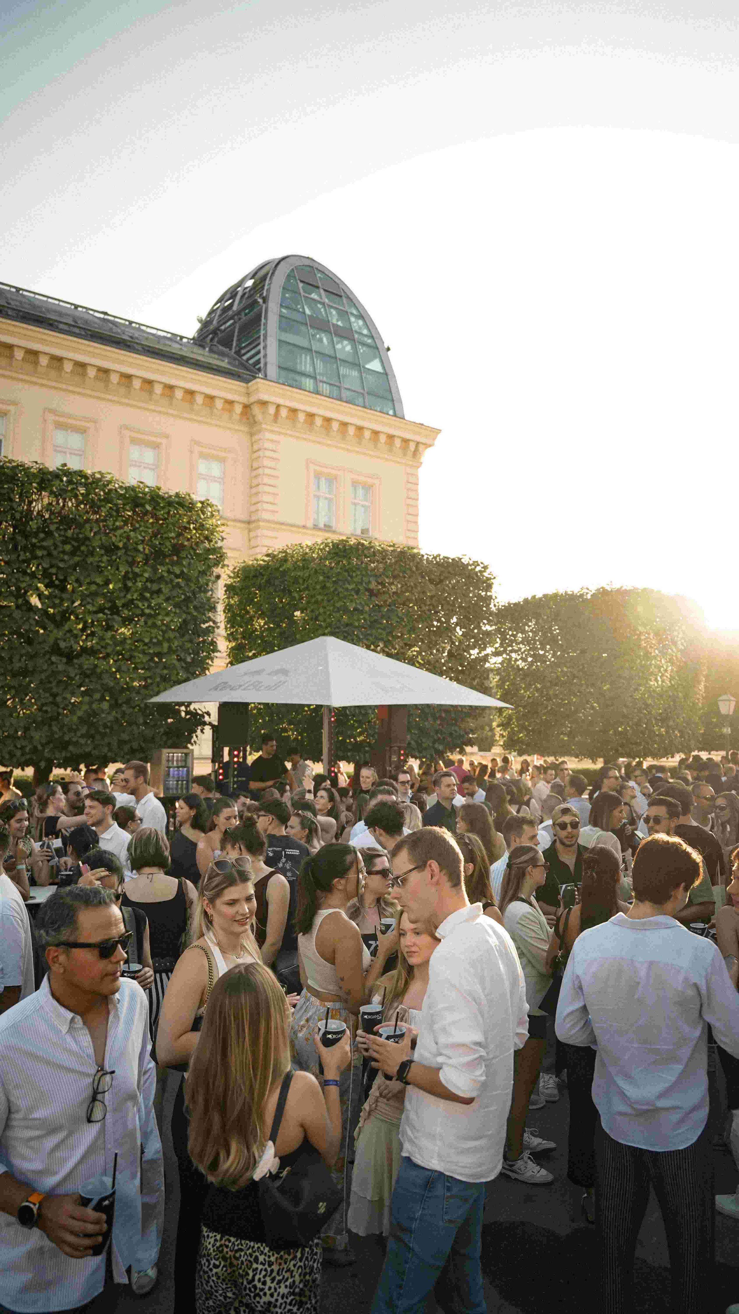 Crowd of people outdoors at a patio with trees and a building with a dome at sunset.