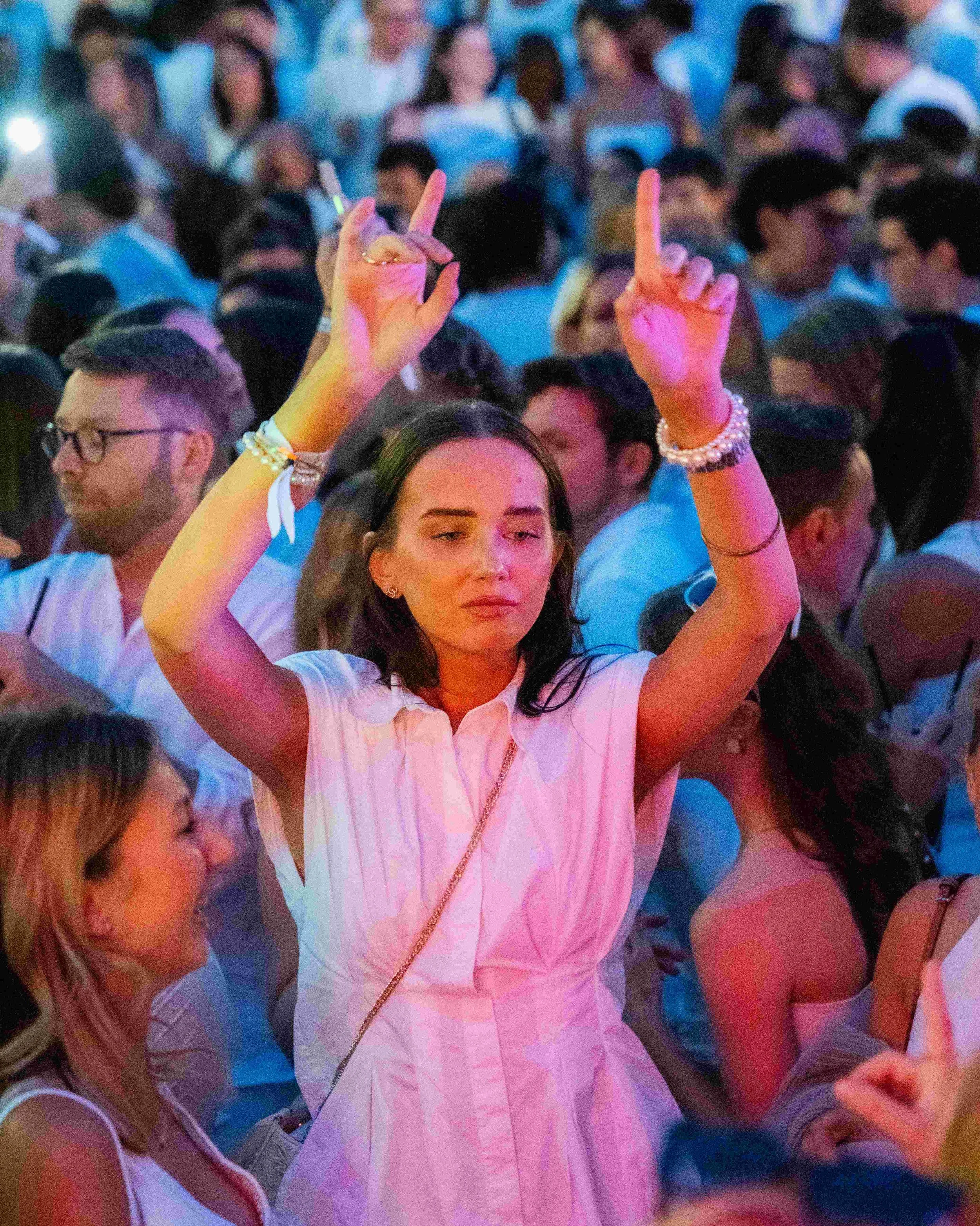 A woman in a white shirt is raising her hands with fingers making a gesture, surrounded by a crowd of people at an event. The crowd appears to be engaged and the scene is illuminated with colorful lighting.