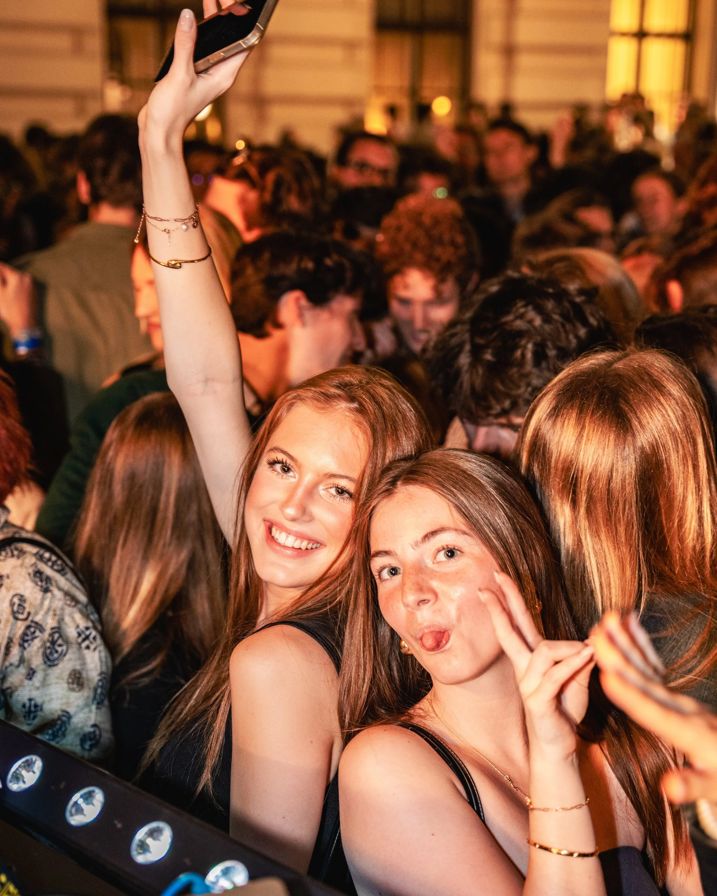 Two young women smiling and taking a selfie at a crowded indoor event or party, with many people in the background.