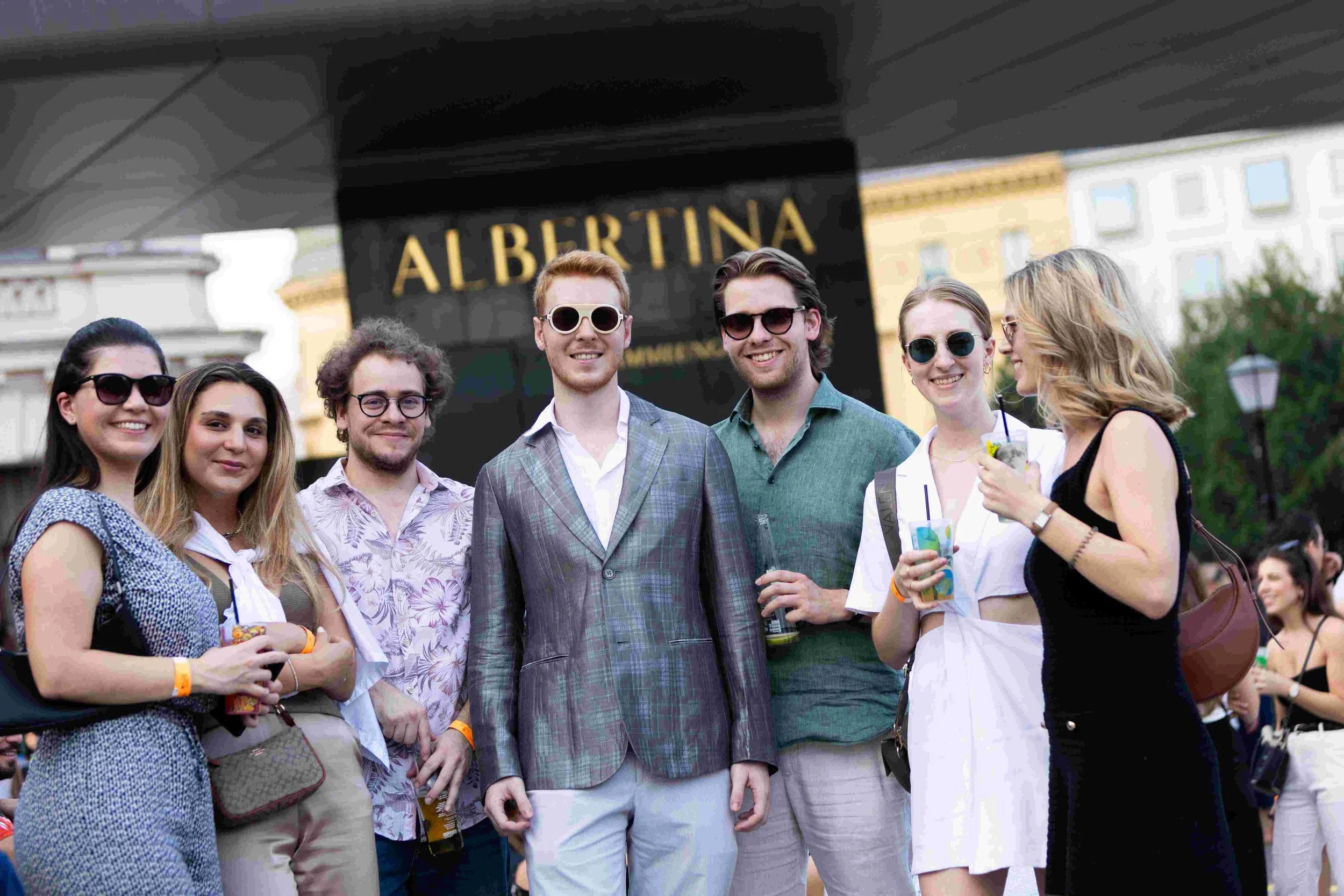 Group of people smiling and posing together outdoors in front of a building with a sign that says "ALBERTINA". They are wearing casual and semi-formal clothing, some with sunglasses, and are holding drinks.