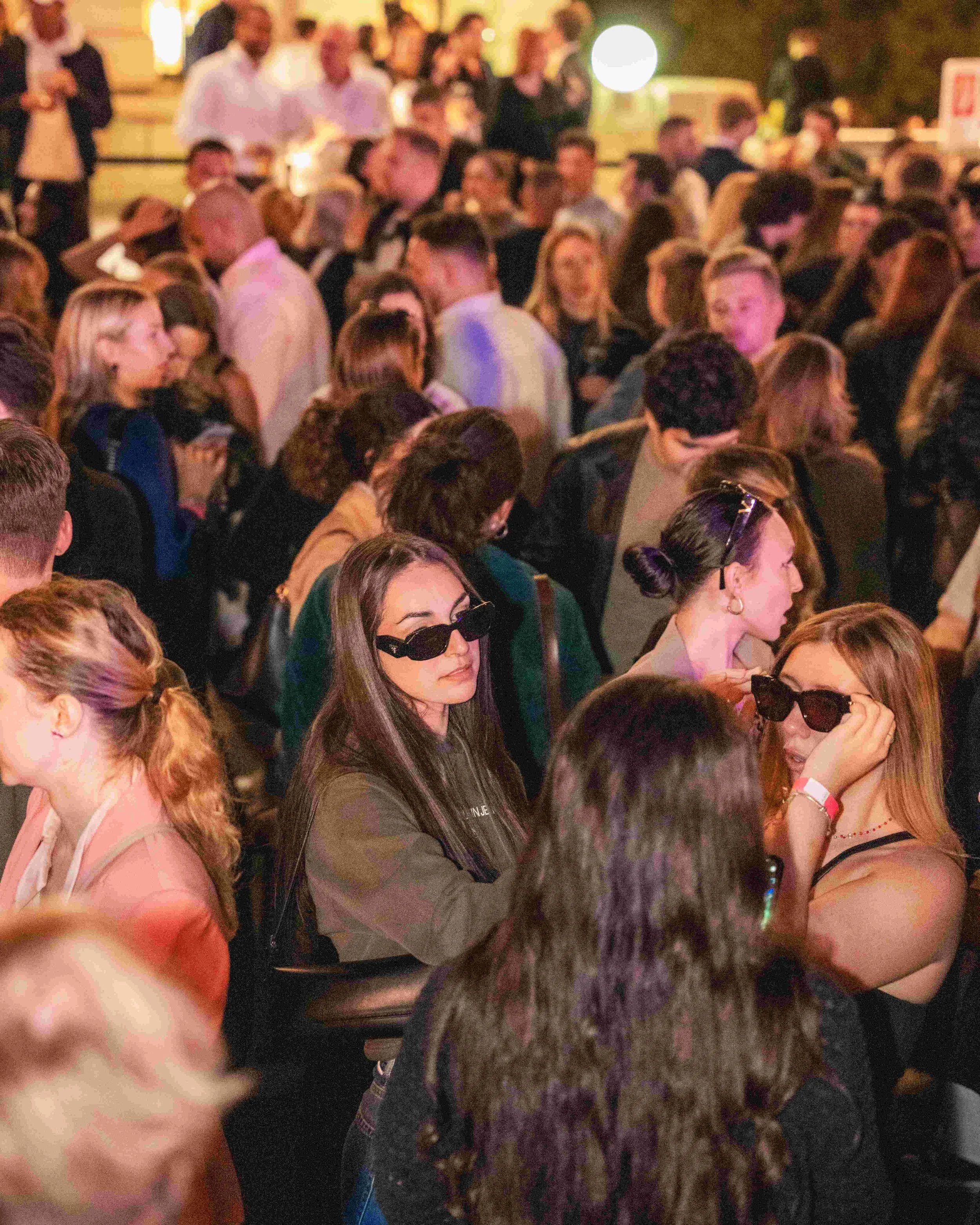 A crowded indoor event with many people, some wearing sunglasses, engaging in conversations.