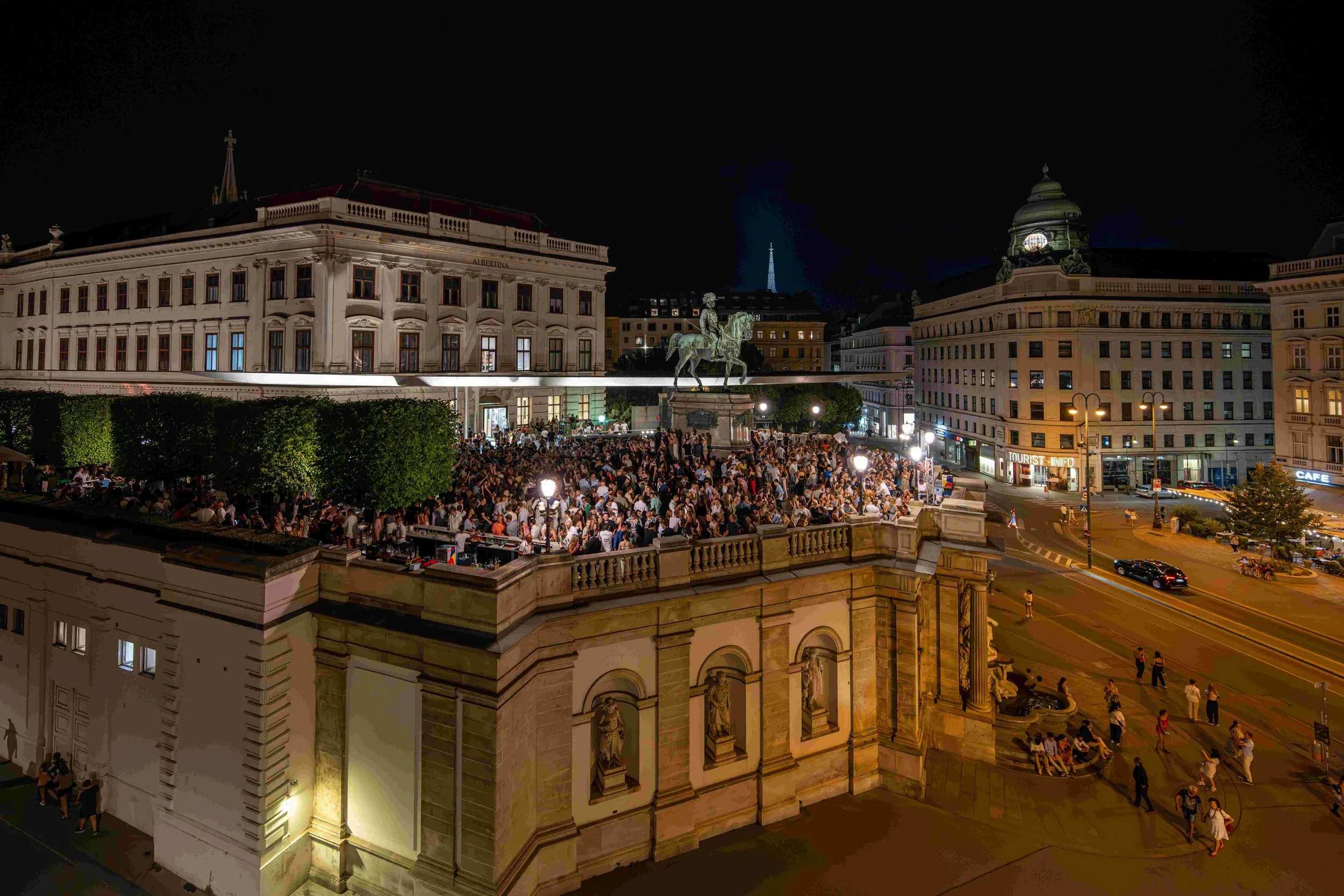Night view of a crowded square with a large equestrian statue, historic buildings, and a crowd of people gathering and sitting.