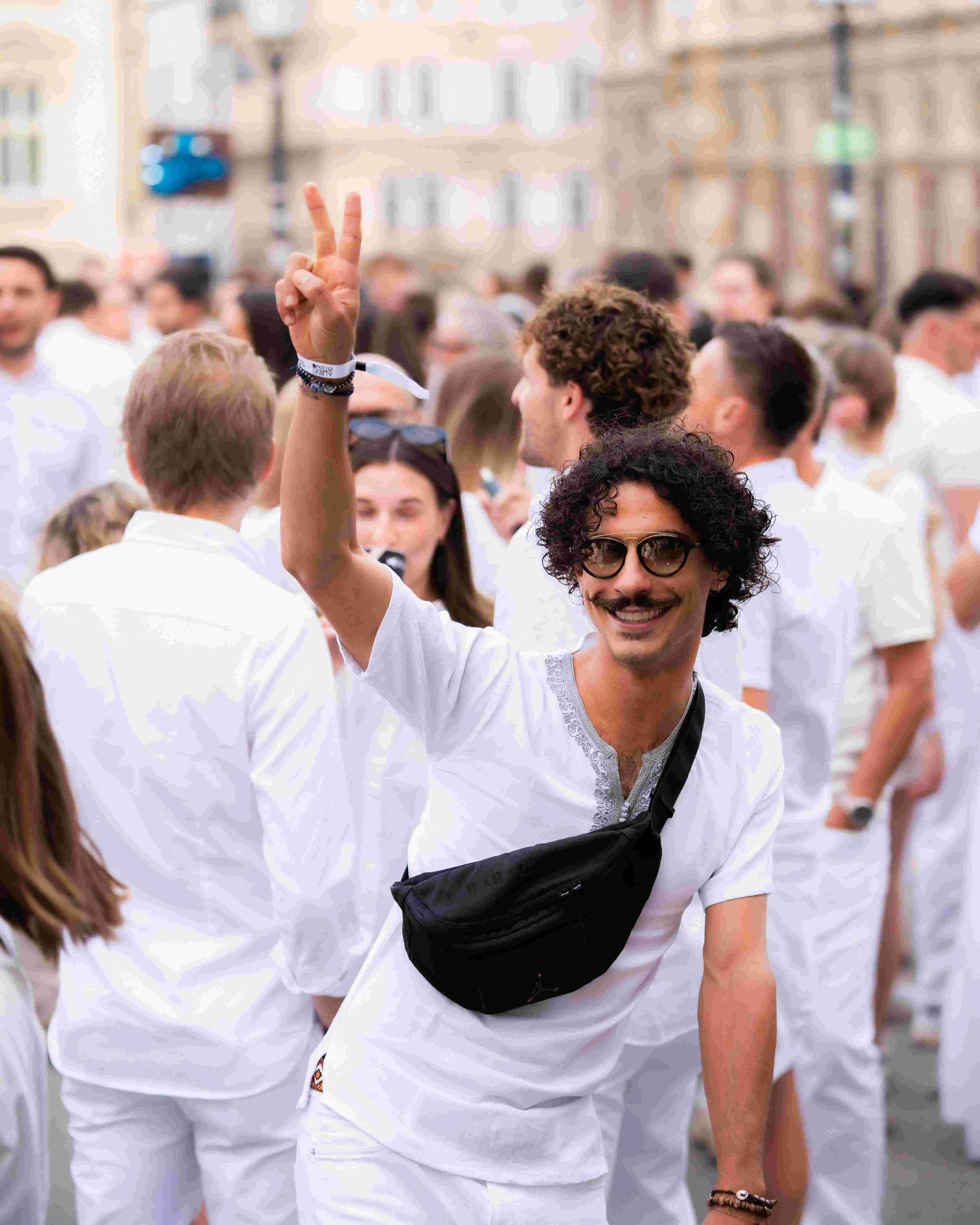 A man with curly hair and sunglasses smiling and raising two fingers in a peace sign at a crowded outdoor event, with people wearing white in the background.