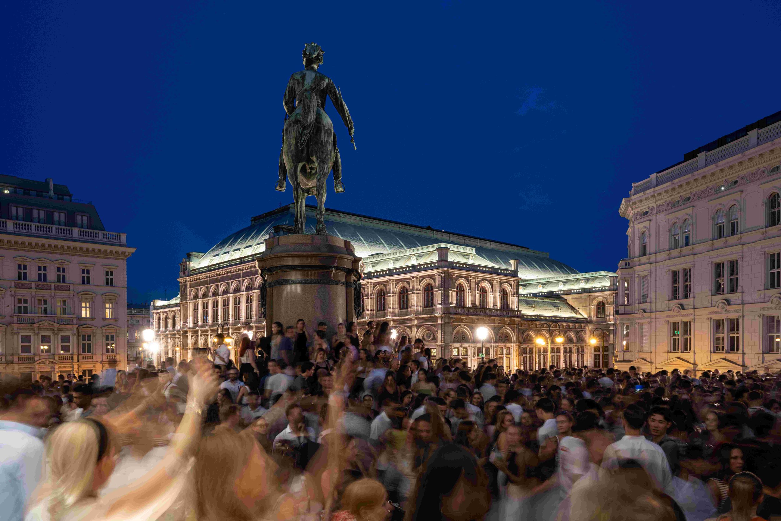 Crowd of people gathered in a city square at night with a large equestrian statue of a woman with a sword on a pedestal in the center, surrounded by historic buildings.