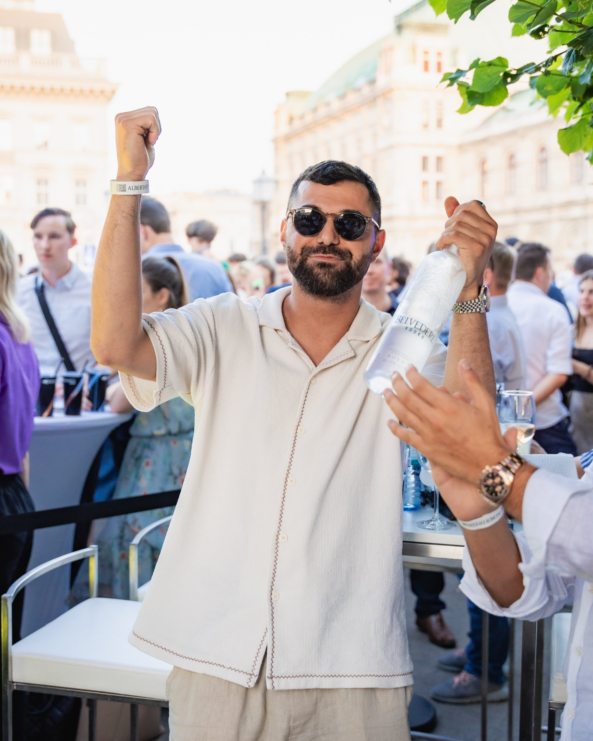 A man wearing sunglasses and a light-colored shirt is raising his right arm in a fist, holding a bottle of Belvedere vodka, at an outdoor event. There are other people in the background and a historic building with a domed roof and ornate architectur