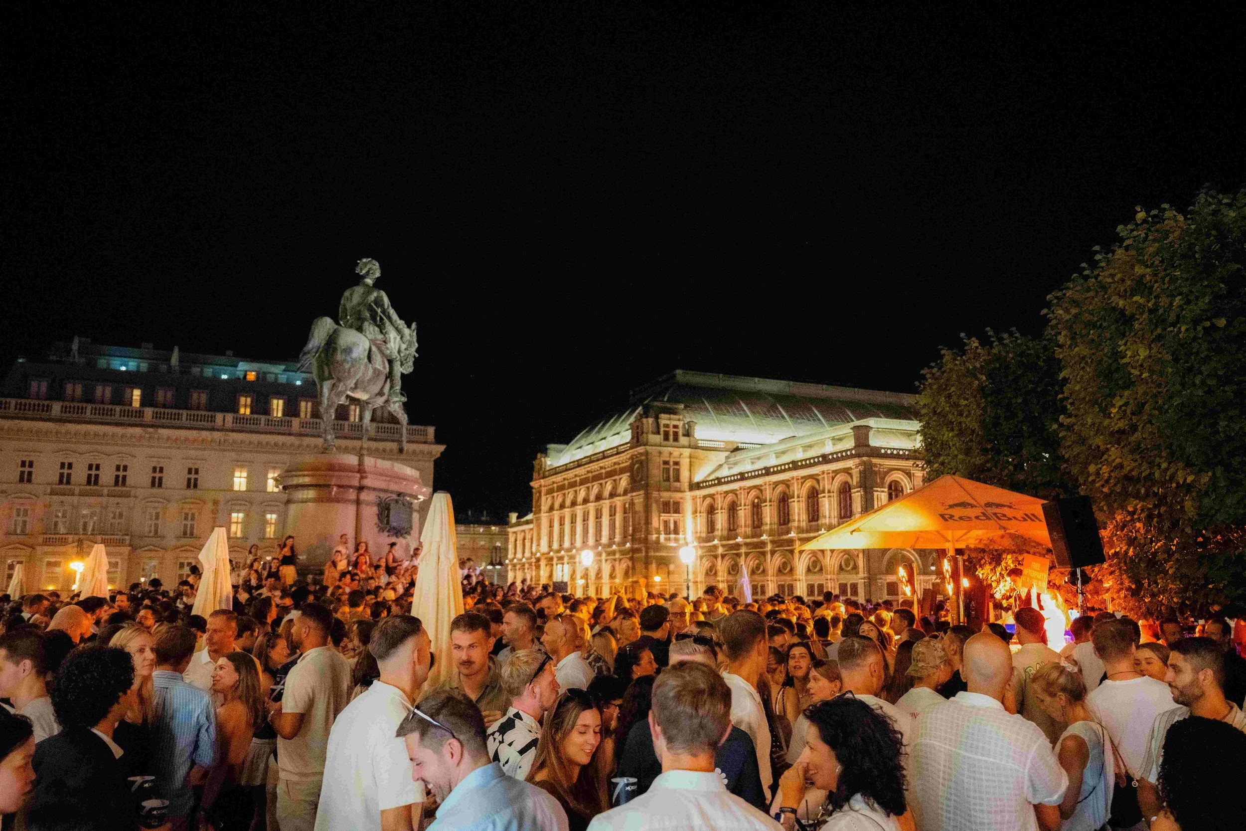 Night scene of a crowded outdoor event with many people, a large statue of a man on horseback, historic building with illuminated windows, and a Red Bull umbrella in the foreground.