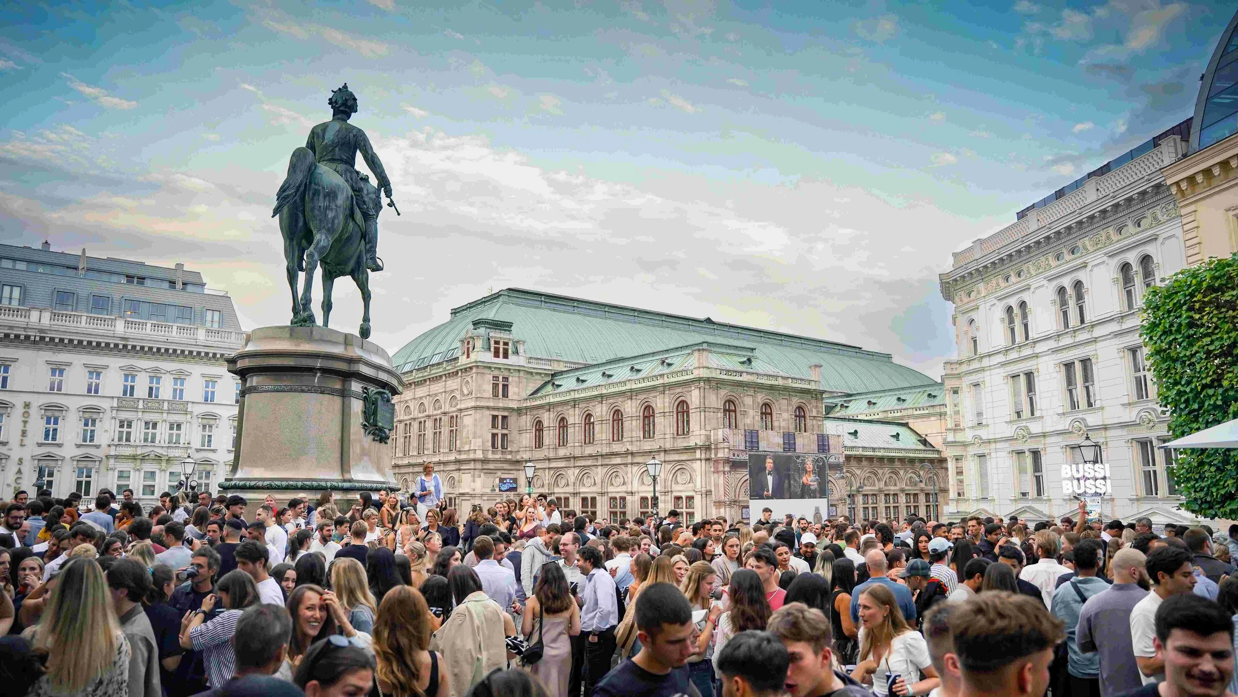 A large crowd of people gathered in a city square with historic buildings, a statue of a man on a horse, and an overcast sky.