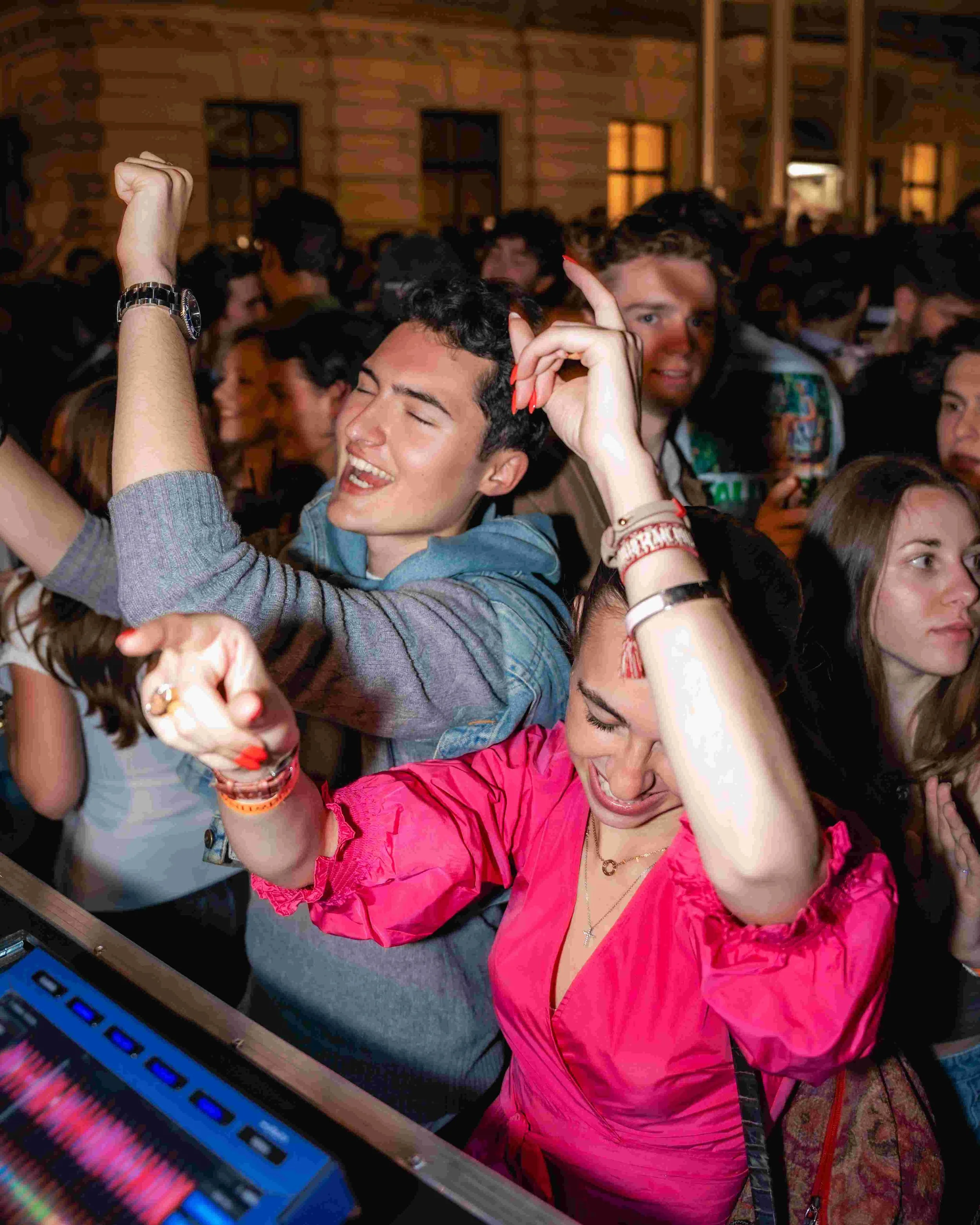 Young woman in pink jacket dancing with eyes closed at crowded indoor event, with DJ equipment visible in foreground.