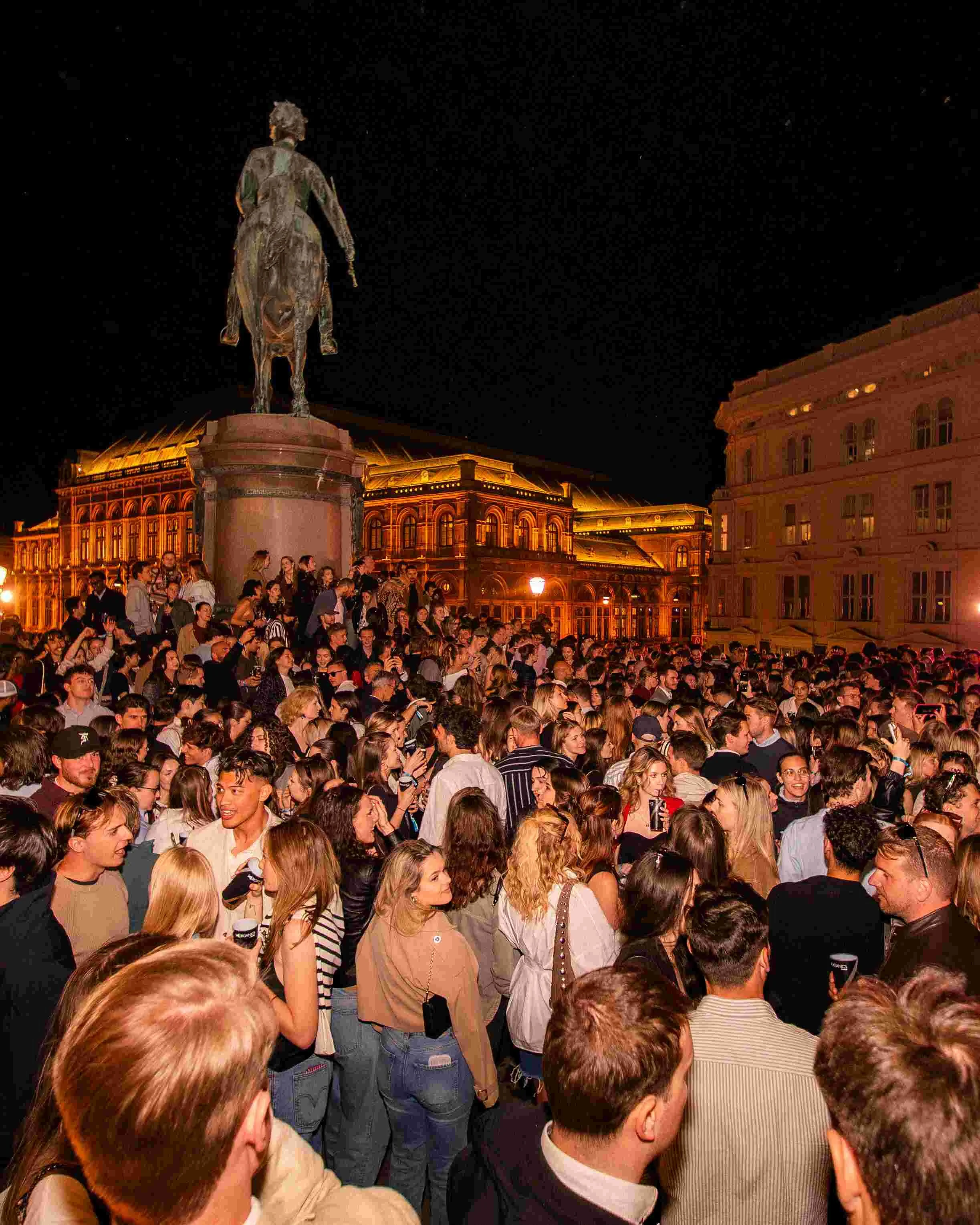 A large crowd of people gathered outdoors at night, looking at a statue of a person mounted on a horse. The building in the background is illuminated with warm lights.