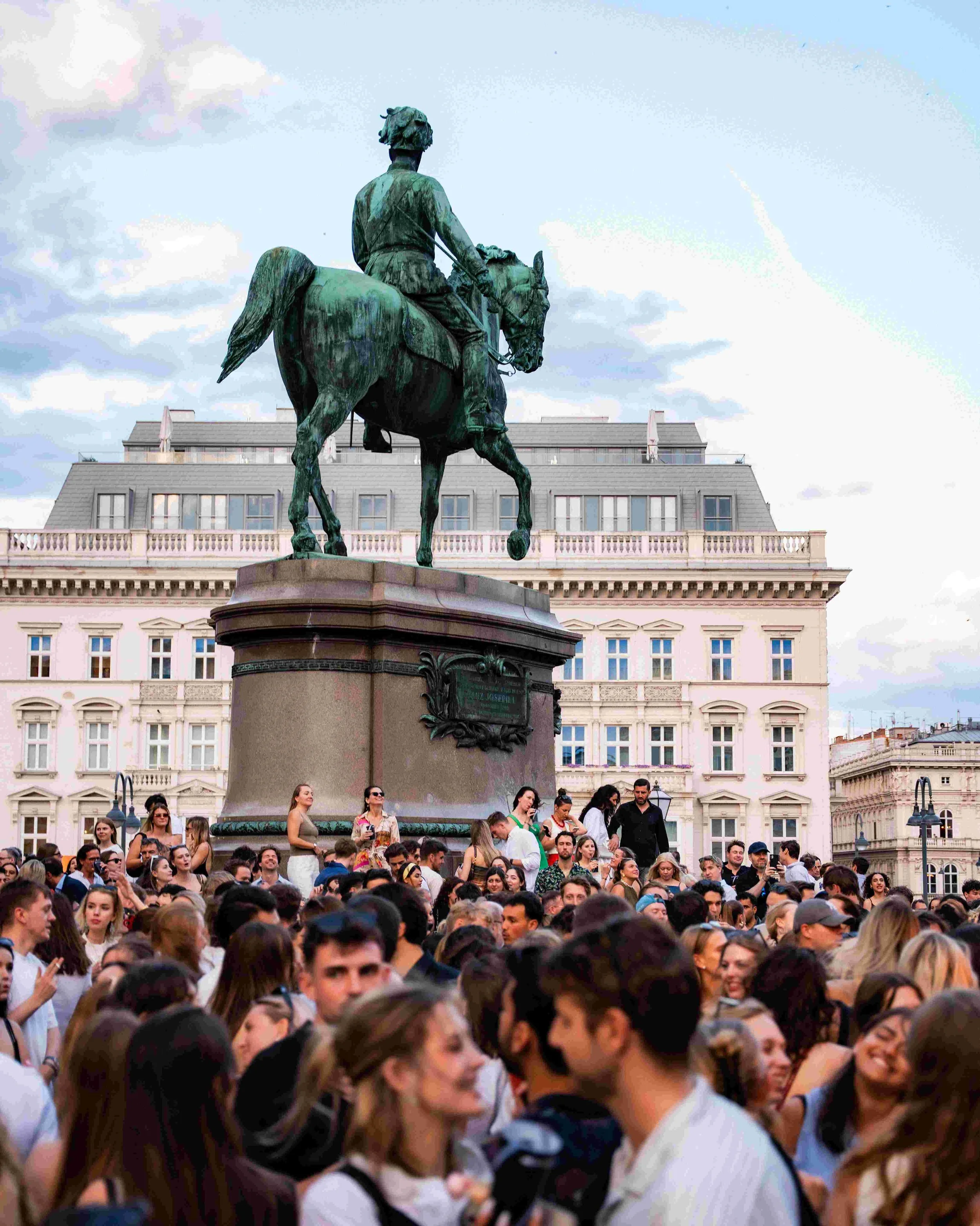 Crowd of people gathered in front of an equestrian statue on a large building. The statue depicts a man on a horse, viewed from behind. The people are of various ages and genders, some looking at phones, others chatting or taking photos. The building
