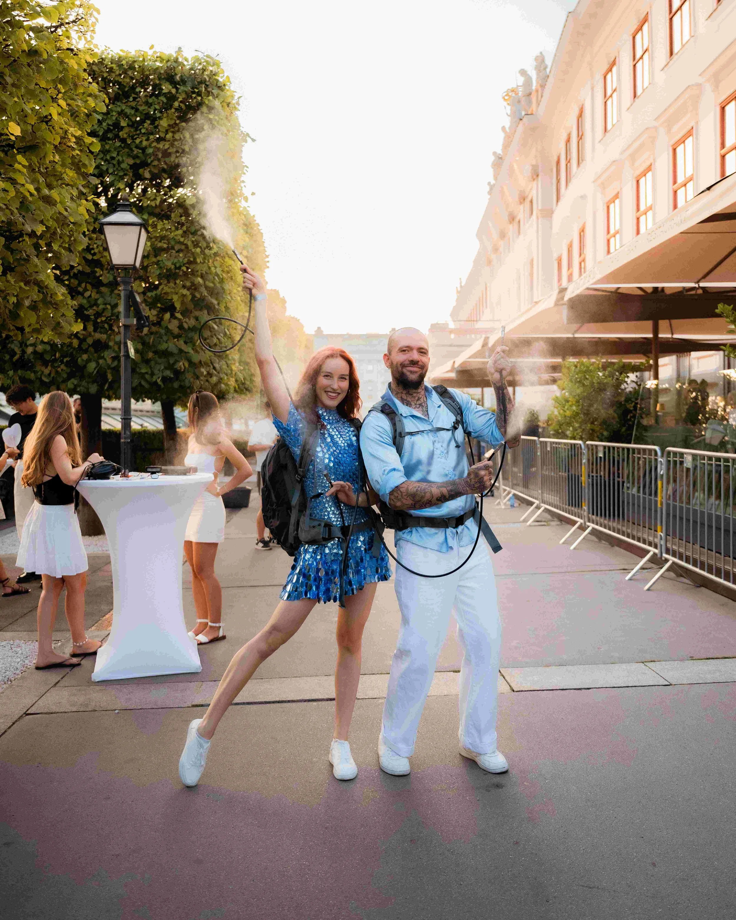 A man and a woman standing together outdoors, both dressed in blue and white, smiling and posing for the camera while holding spray bottles. The woman is wearing a shiny blue dress and white shoes, and the man is wearing white pants and a light blue 