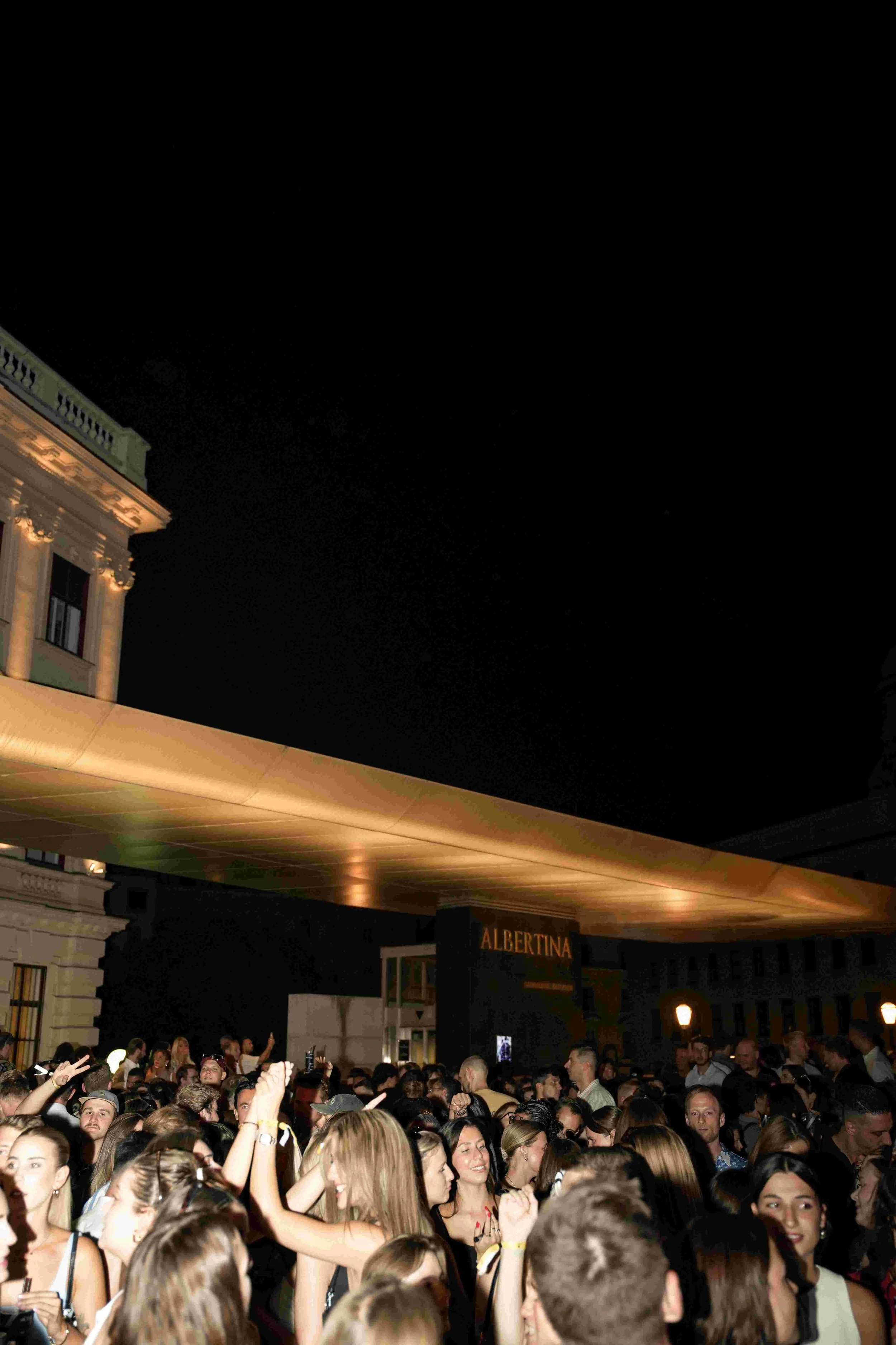 A large crowd of people at a nighttime outdoor event, with a building and a modern canopy overhead, with some people dancing and taking photos.