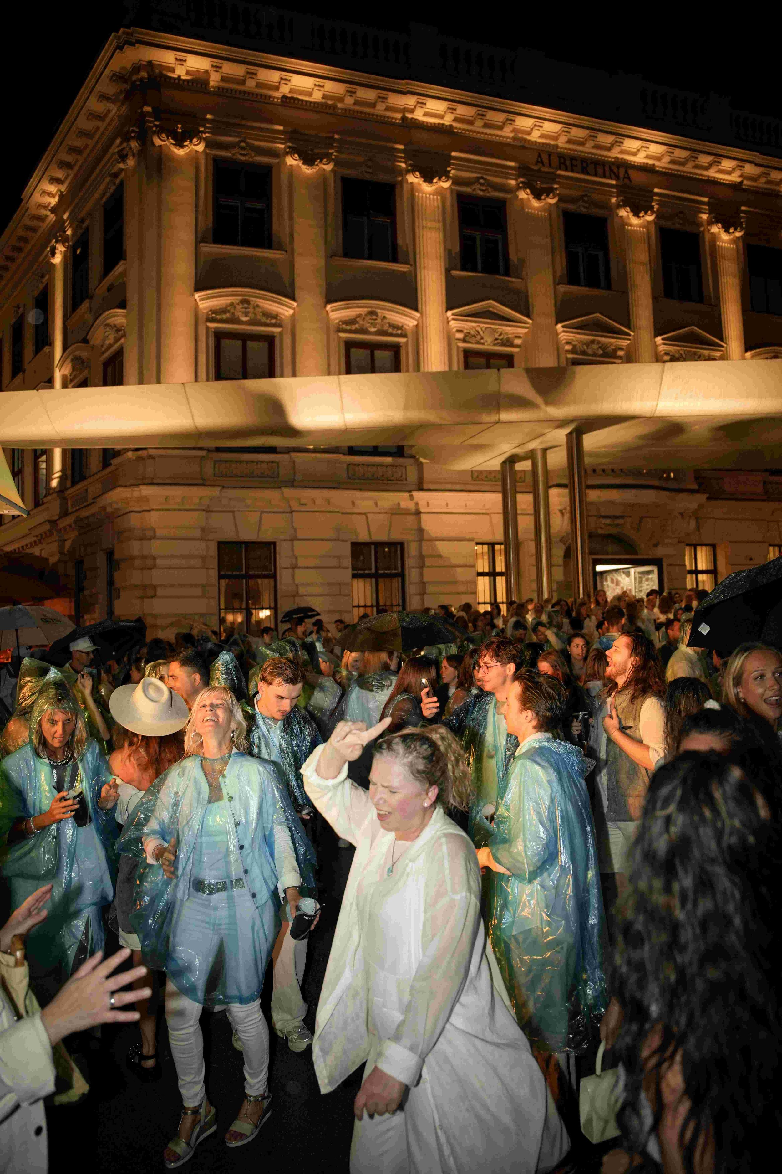 Crowd of people gathered outside a lit building at night, many wearing rain ponchos, some holding umbrellas, amidst a lively atmosphere.