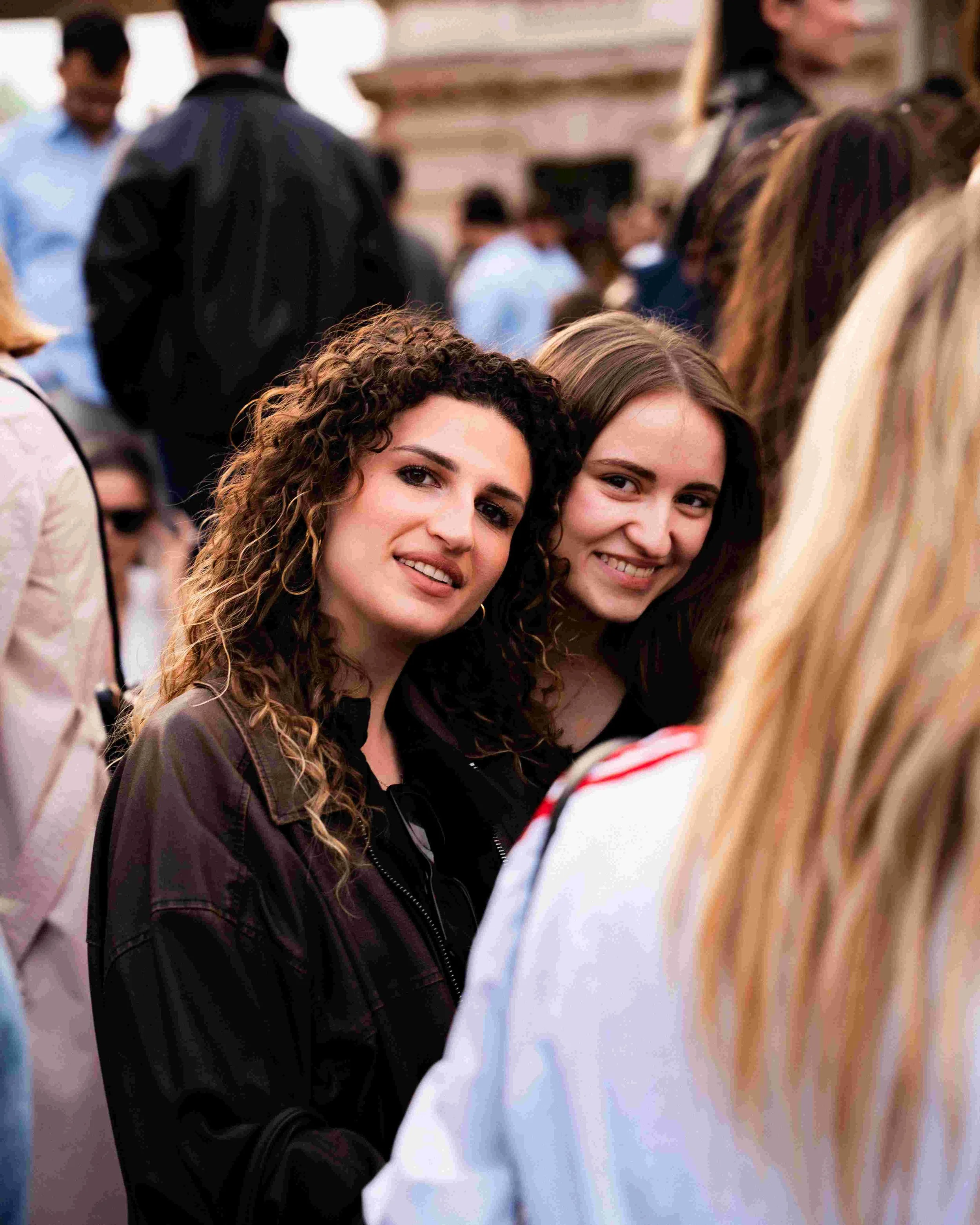 Two young women with curly and straight brown hair smiling and looking at the camera, with blurred people in the background at an outdoor event.