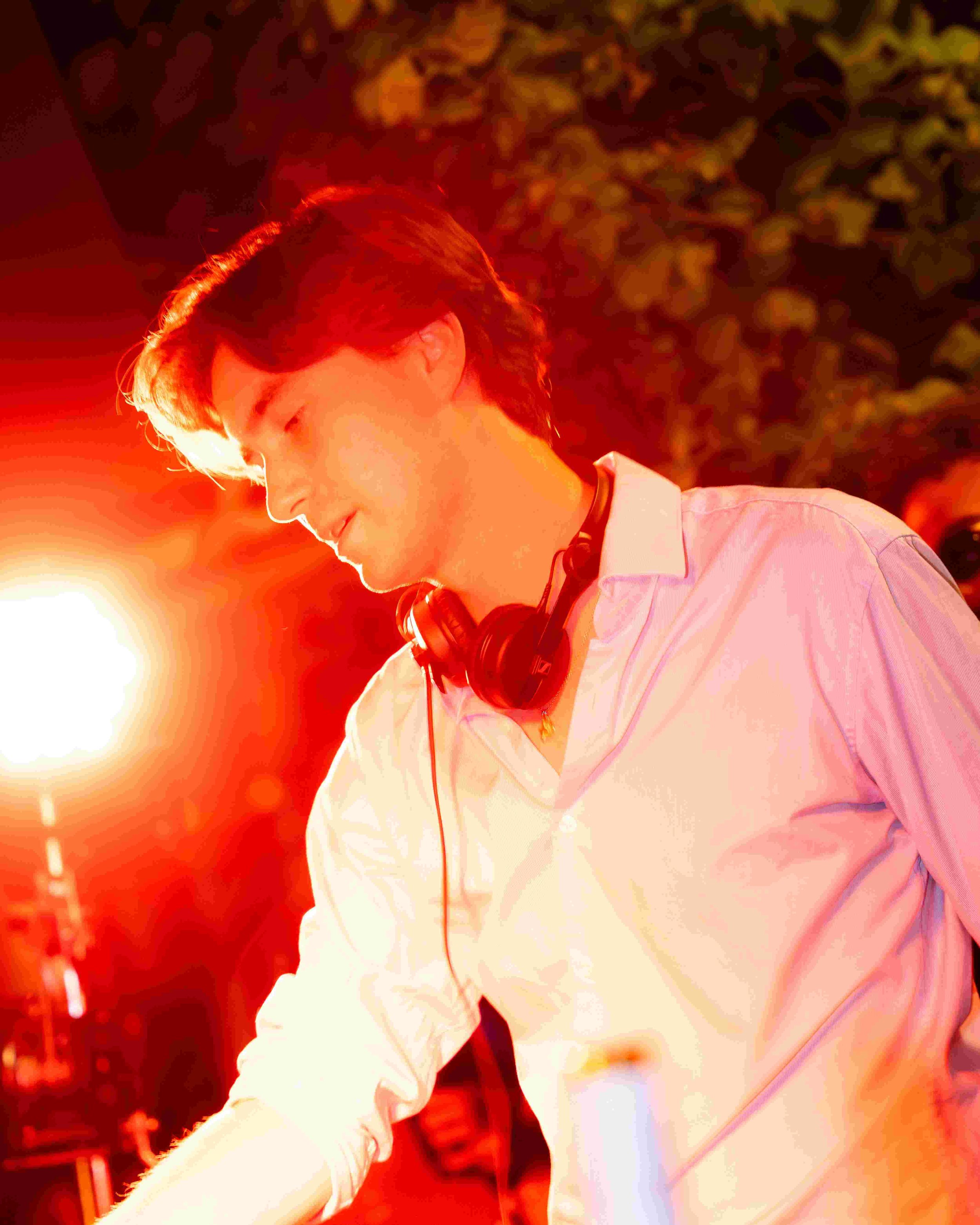 A young man with headphones around his neck, wearing a light-colored shirt, looking downward, with a warm orange glow in the background.