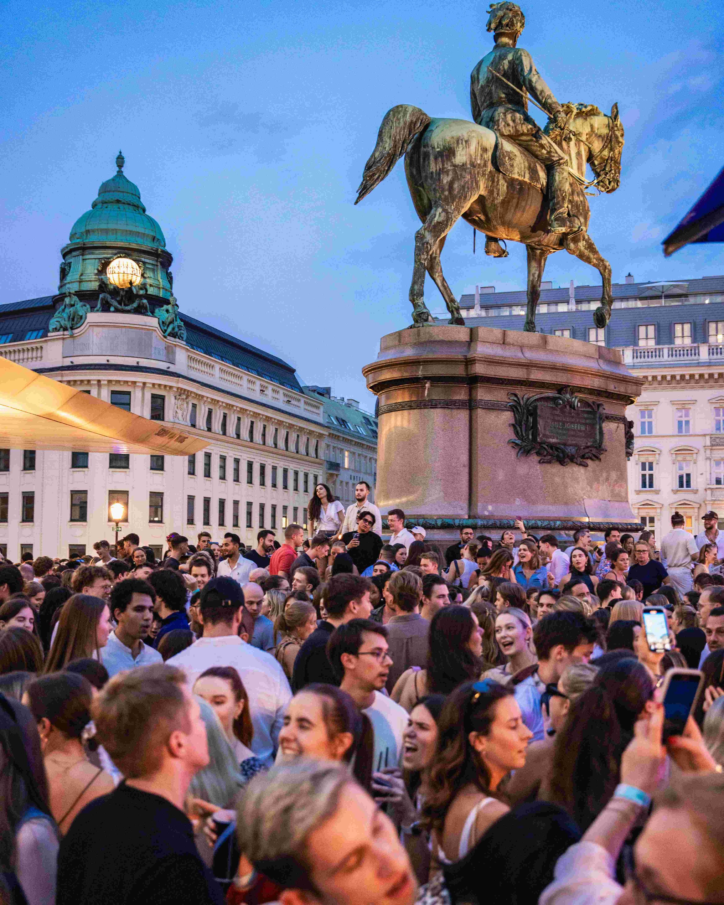 A large crowd of people gathered outdoors in an urban setting during evening, with an equestrian statue and historic buildings in the background.