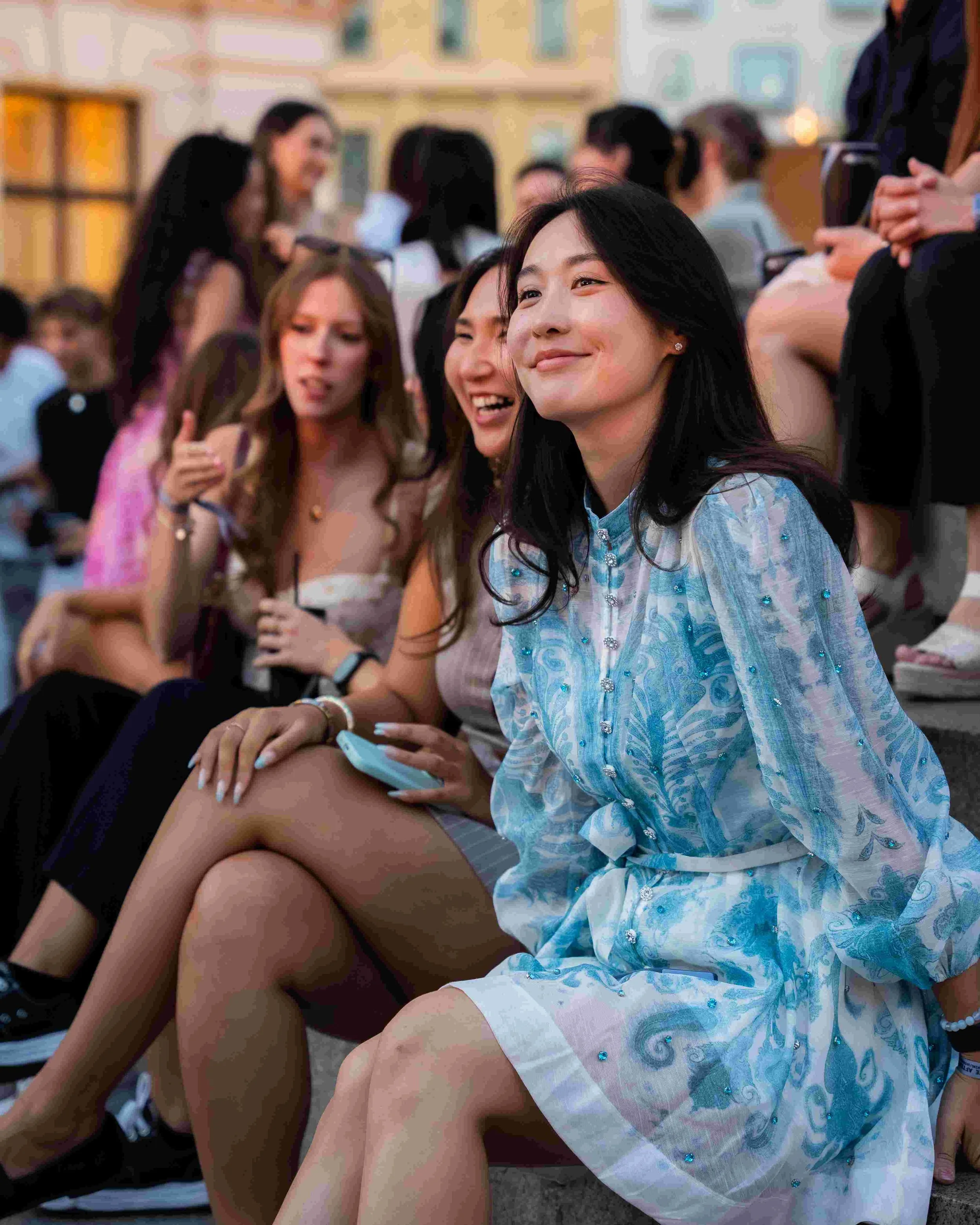 A group of women sitting outdoors, smiling and chatting, some holding phones. One woman in the foreground is dressed in a light blue patterned dress, smiling. The background shows more women engaged in conversation, with a building and decorative lig