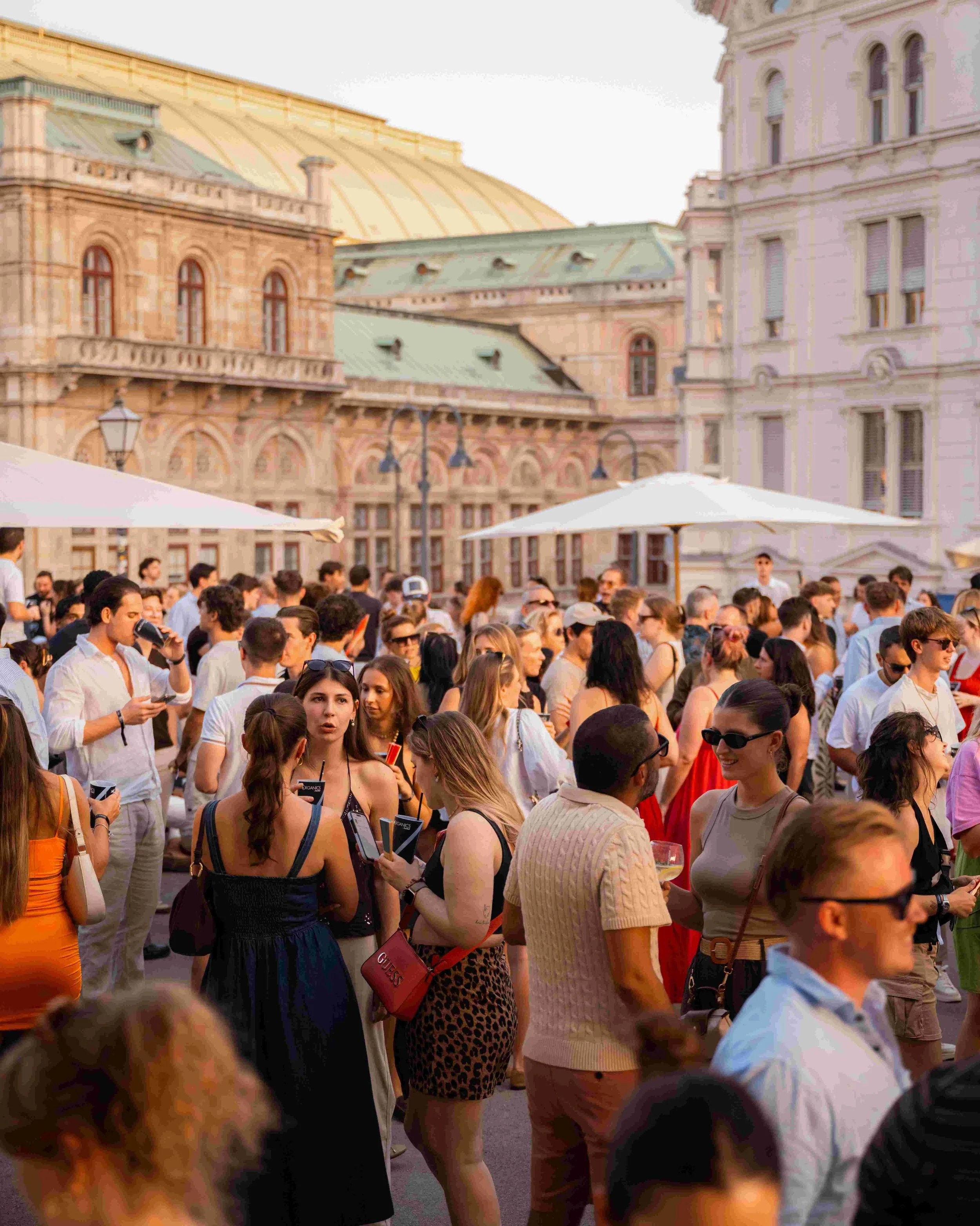 A large crowd of people gathered outdoors in front of historic buildings with ornate architecture, under umbrellas on a warm sunny day.