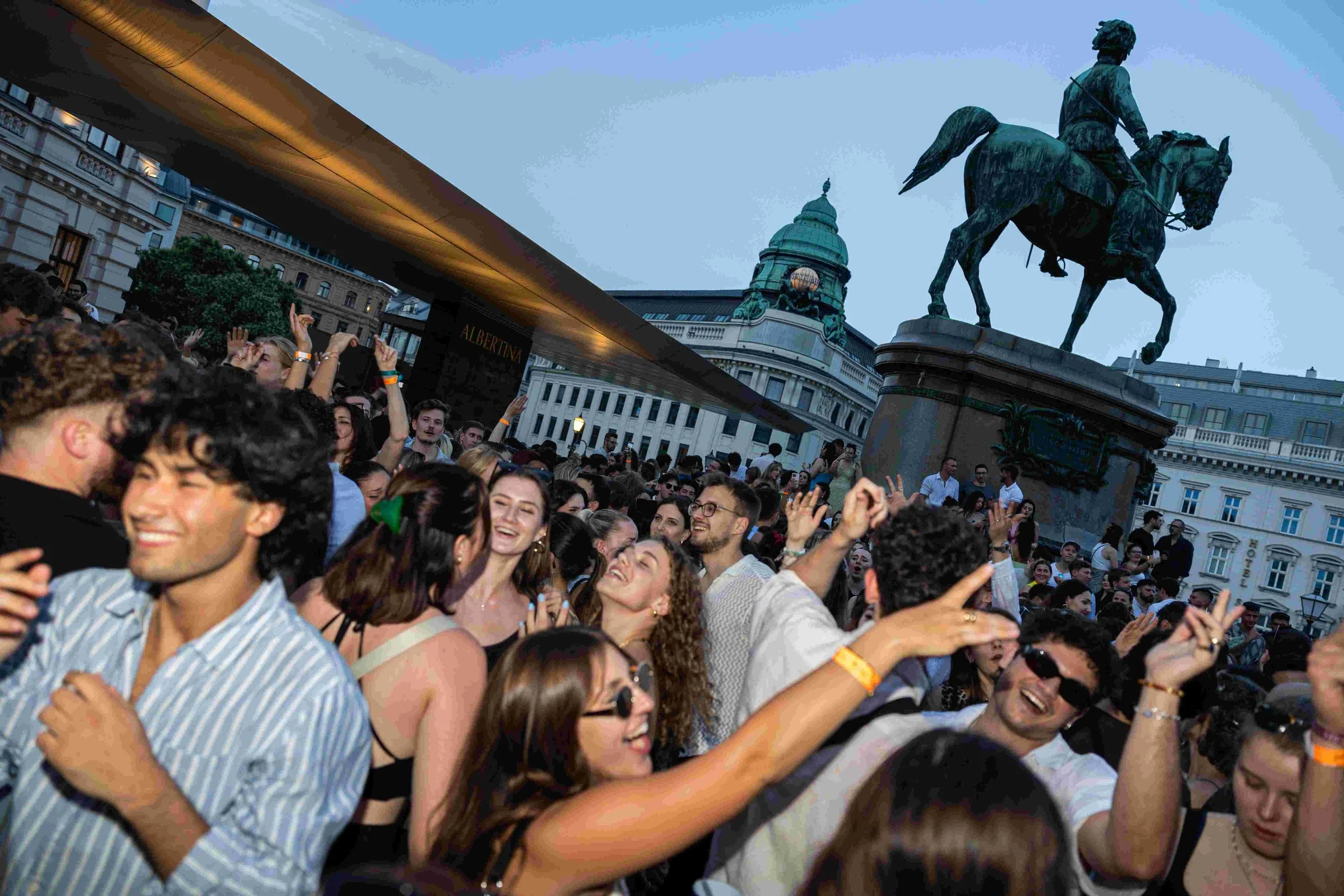 A large crowd of young people gathered in a city square, smiling and dancing, with an equestrian statue and historic buildings in the background during twilight.
