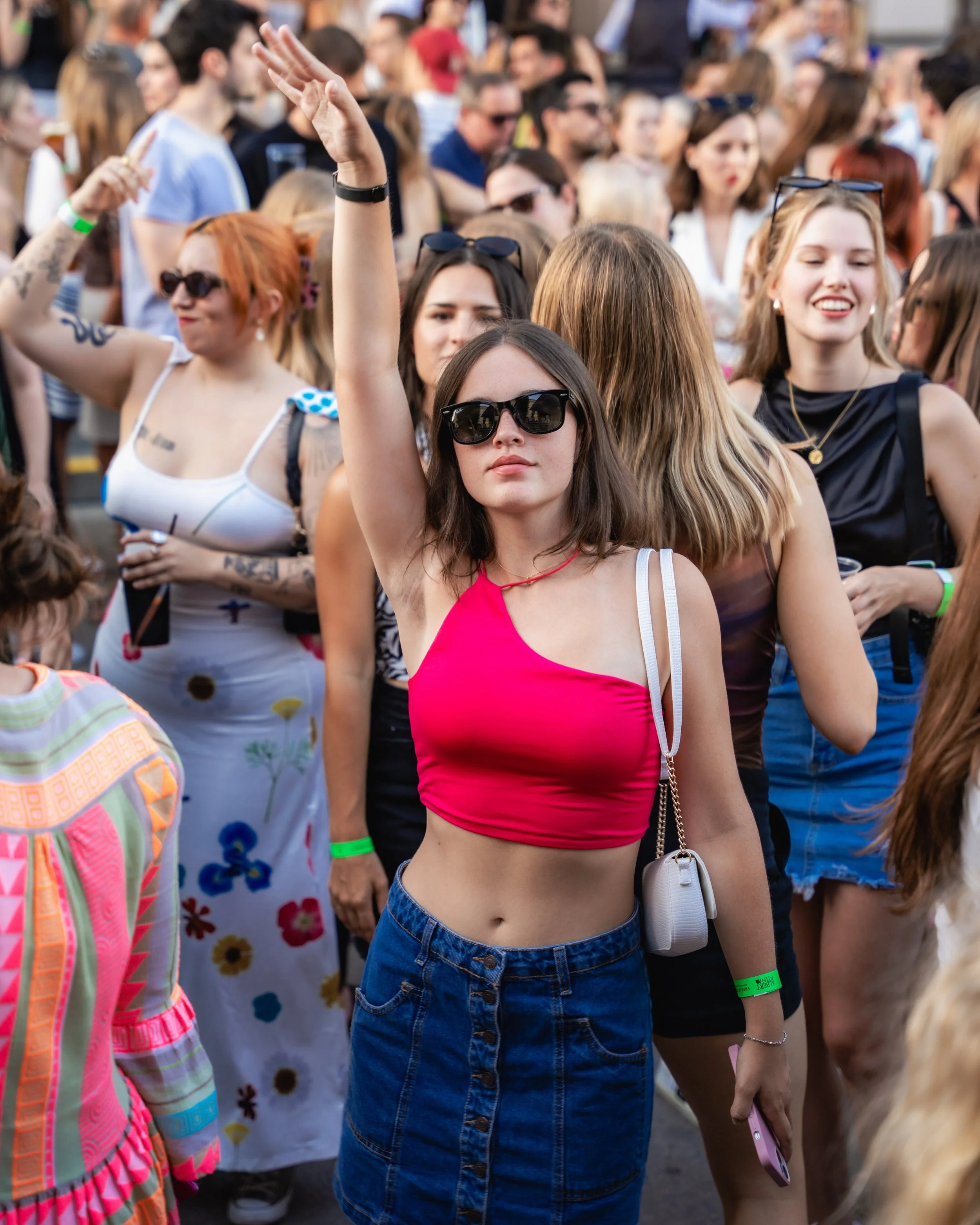 Young woman with long brown hair, wearing sunglasses, a pink tank top, and high-waisted jeans, standing in a crowd at an outdoor event, holding a pink phone, with her arm raised.