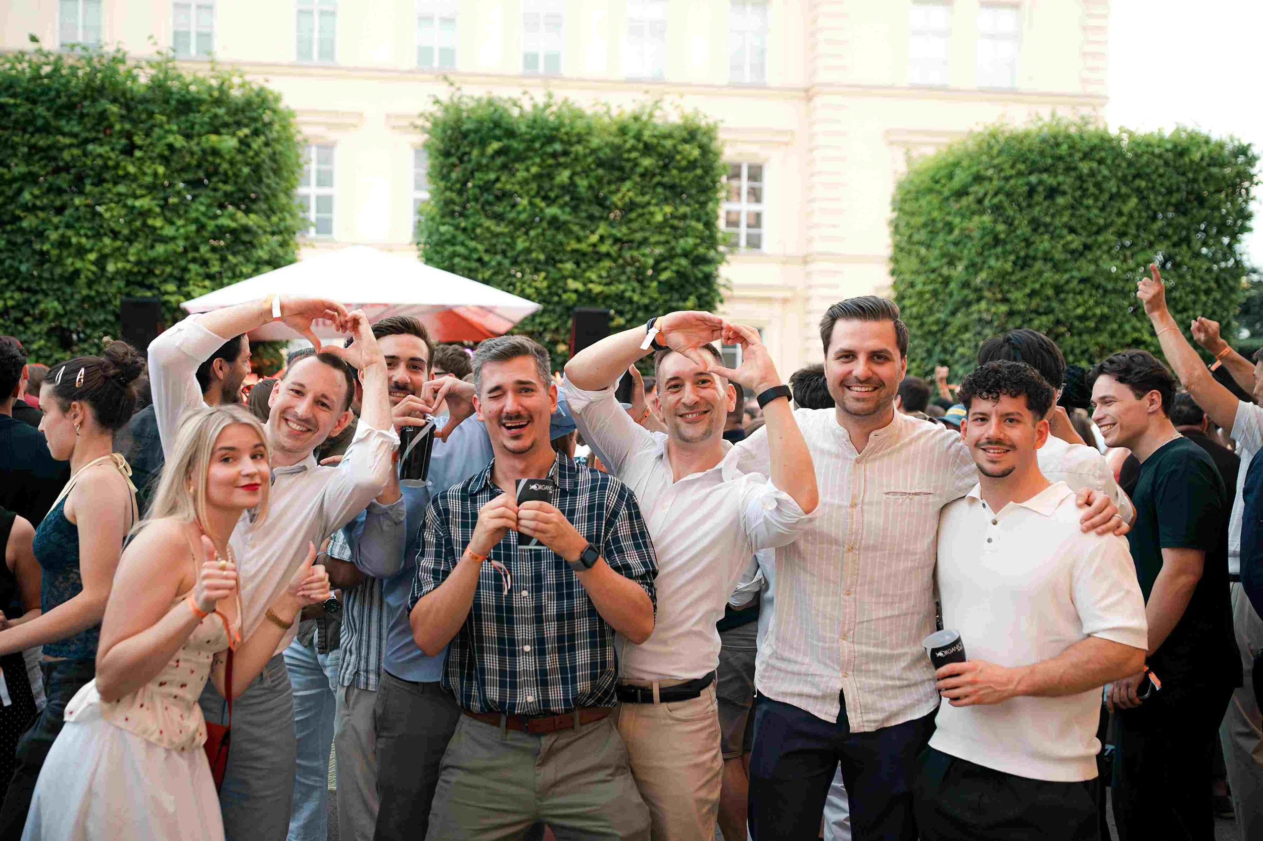 A large group of diverse young adults at an outdoor event, smiling and making heart shapes with their hands. Some are holding drinks. There are trees and a building in the background.