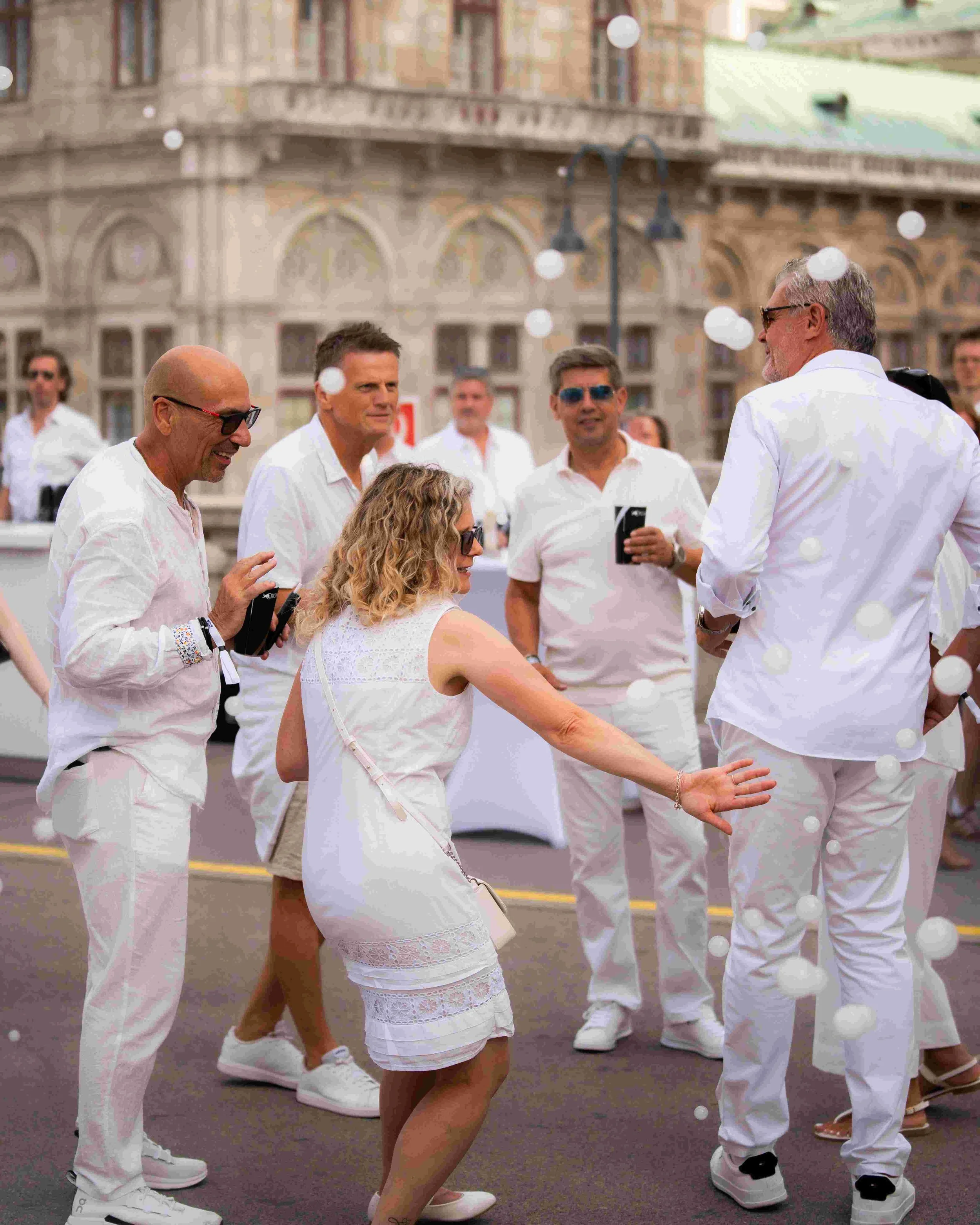 Group of people dressed in white clothing, socializing outdoors with historic building in the background.
