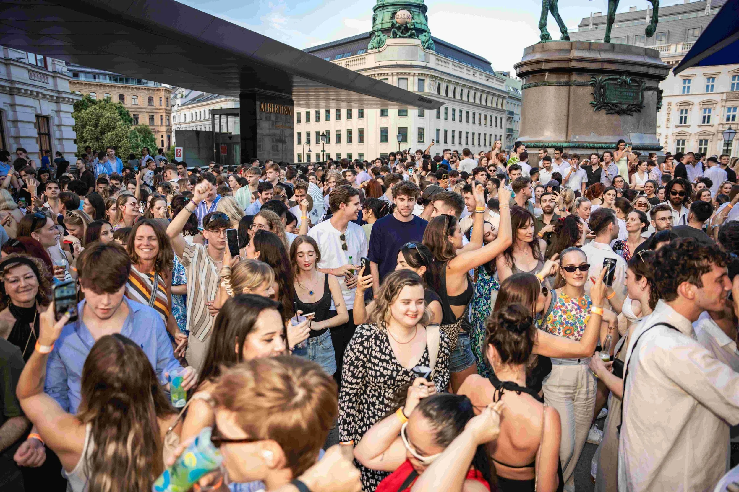 A large crowd of people gathered outdoors in a city square, many holding up their phones, with historic and modern buildings and statues in the background.