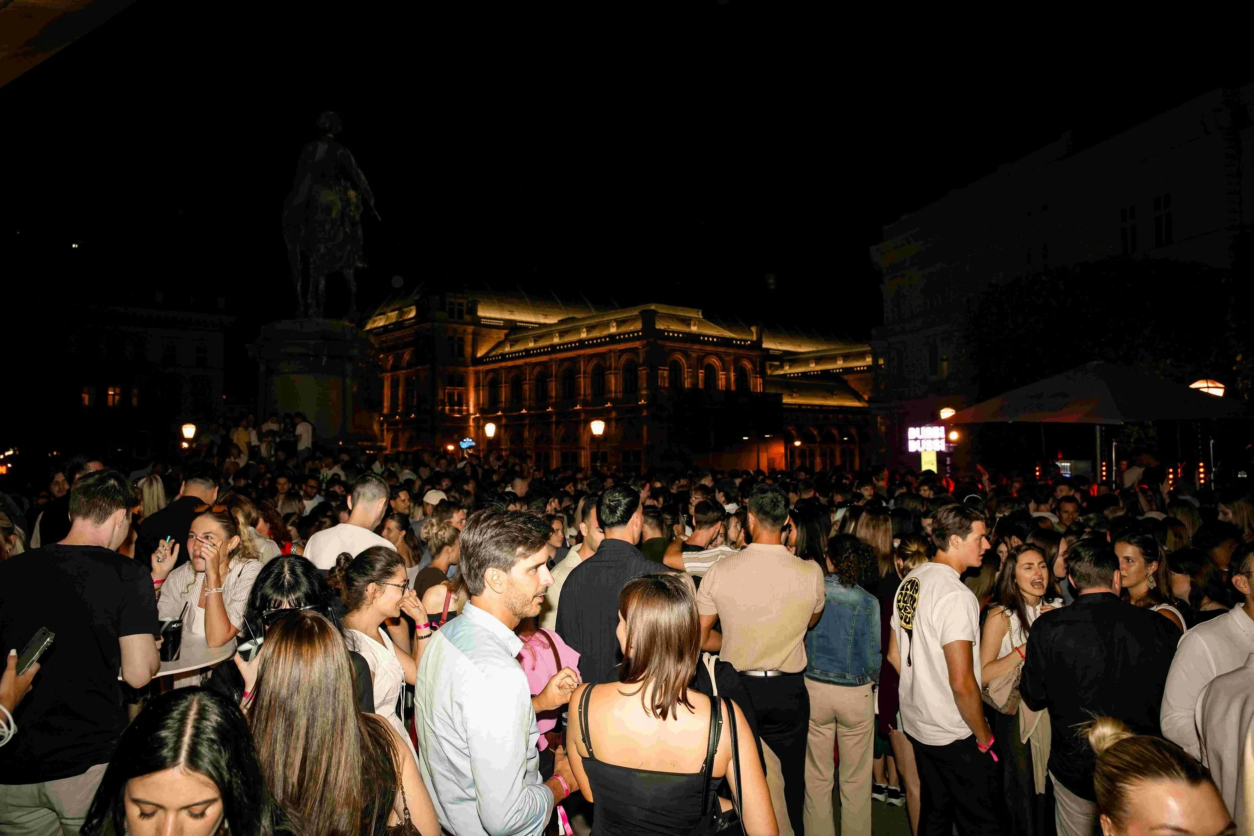 A large crowd of people gathered outdoors at night, with a historic building illuminated in the background and a statue of a person standing atop a pedestal.