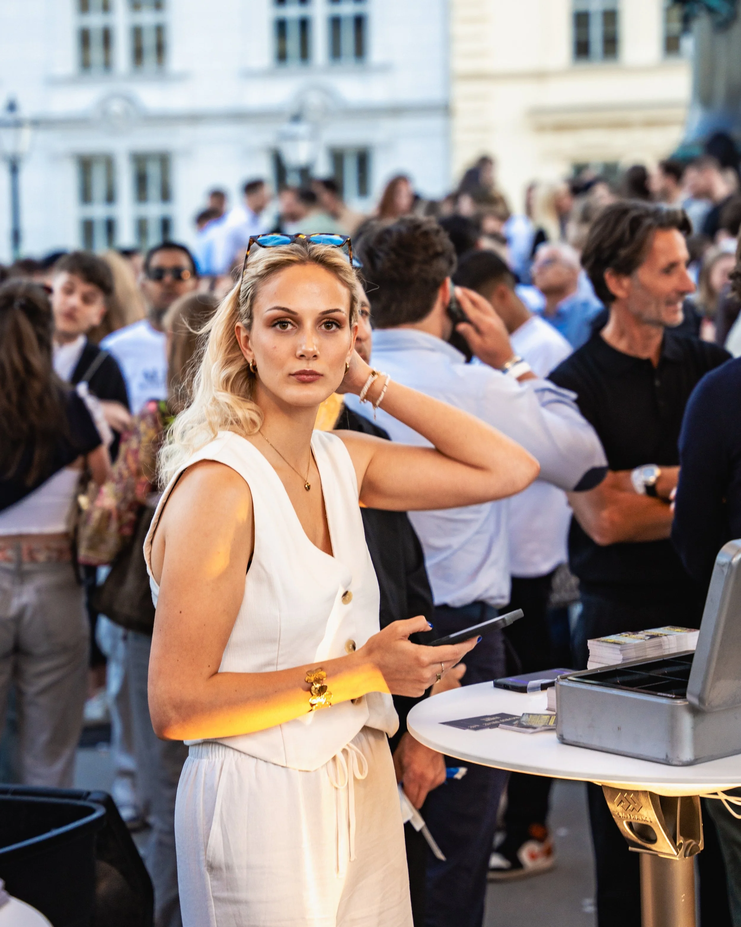 A young woman with blonde hair, sunglasses on her head, gold jewelry, and a white sleeveless dress holding a smartphone at an outdoor event with a crowd in the background.