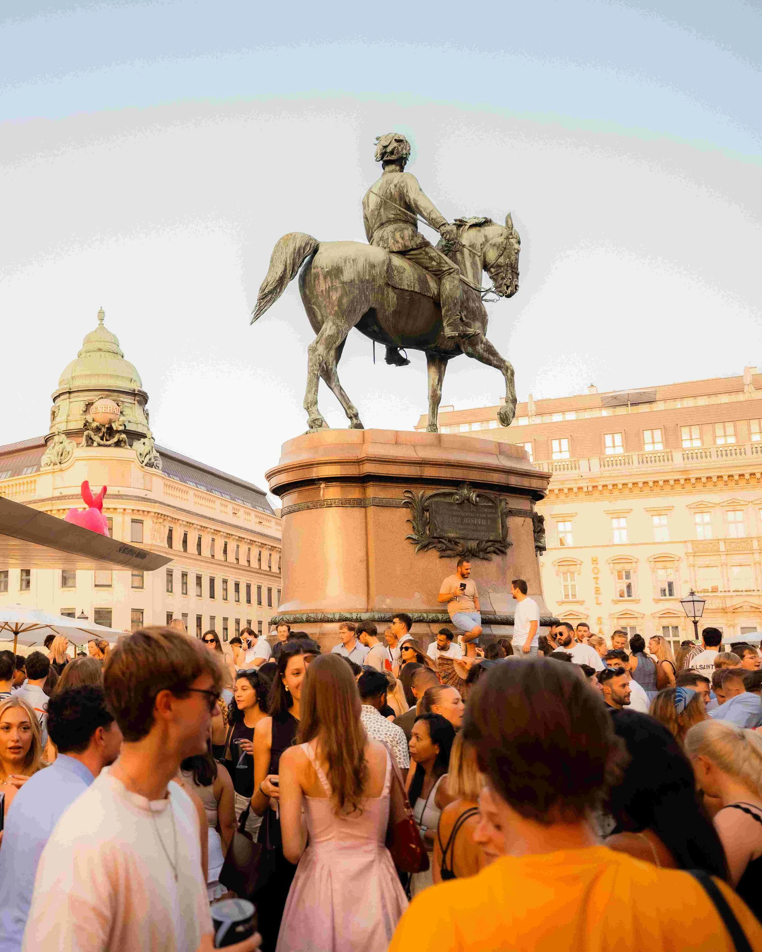 Crowd of people gathered around a large statue of a man on a horse in a city square during daylight.