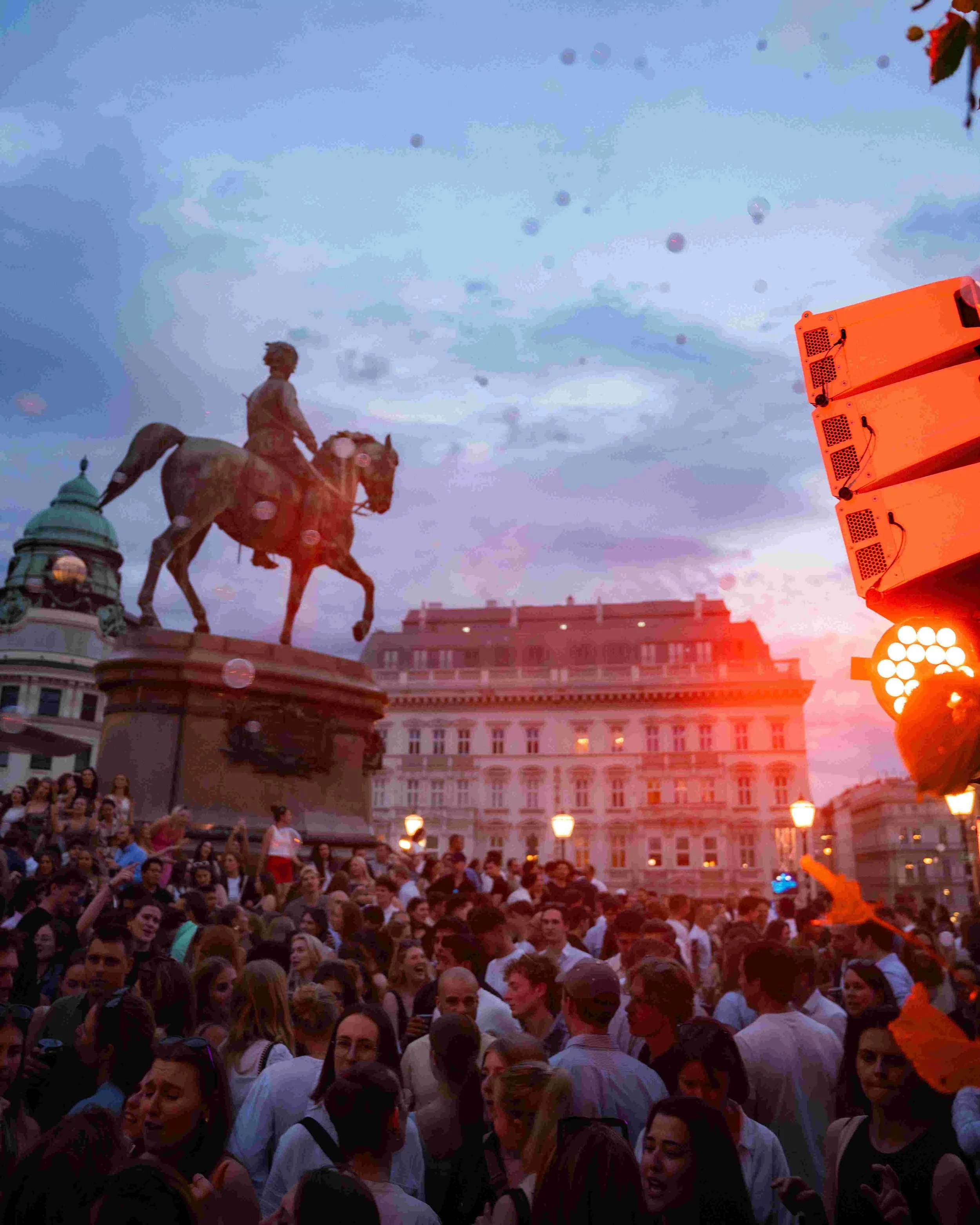 Crowd gathered at an outdoor event during the evening, with statues of a man on a horse and a woman, surrounded by historic buildings and a large orange speaker.