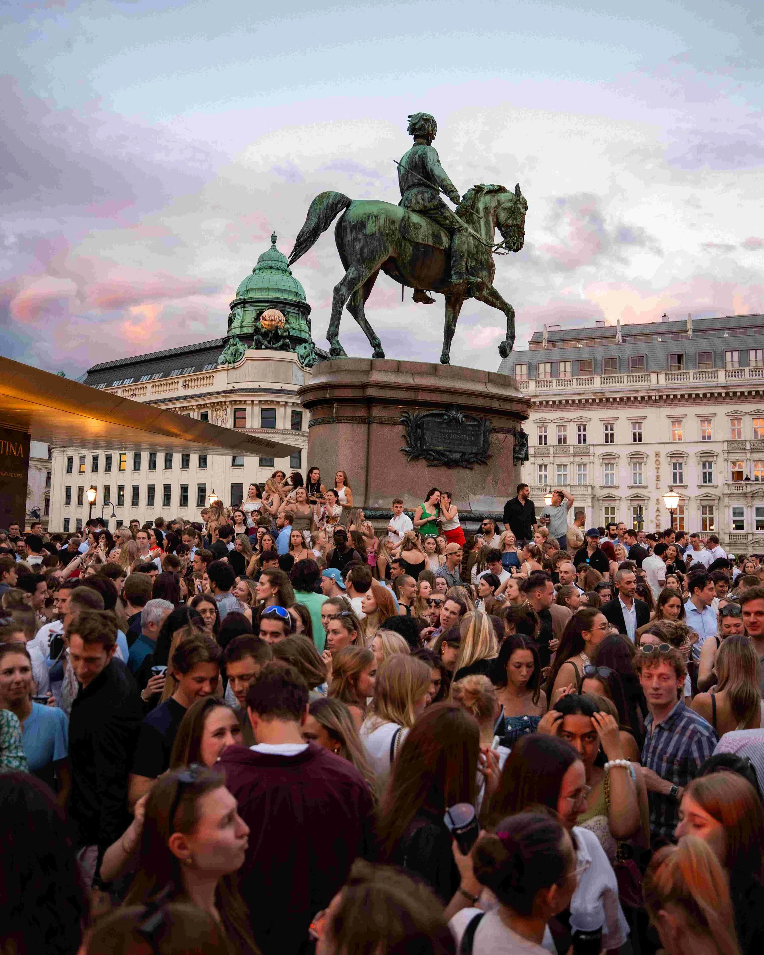 Crowd of people gathered at an outdoor event in a city square, with a large equestrian statue of a historical figure in the background, under a partly cloudy sky during sunset.