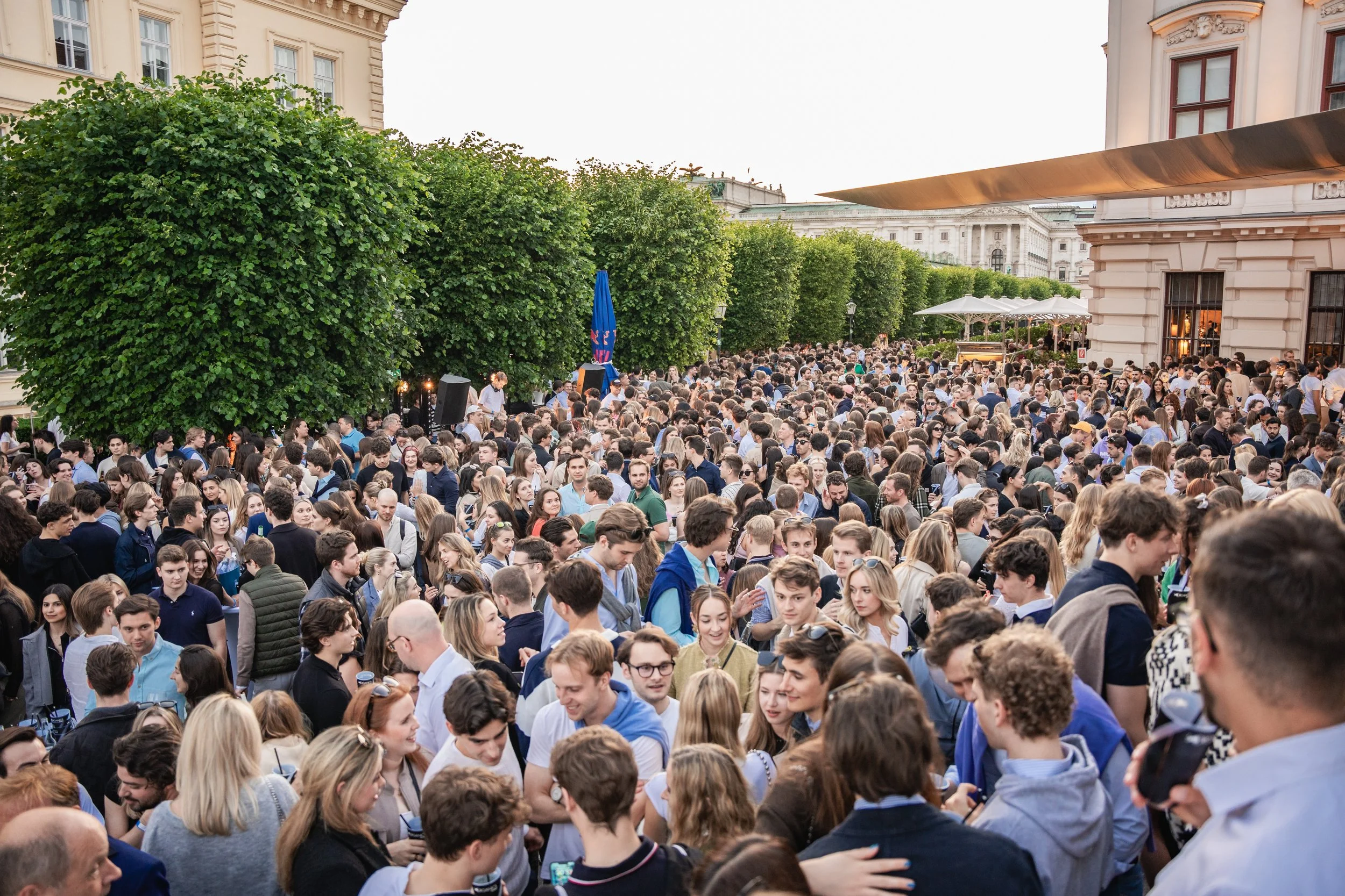 A large crowd of people gathered outdoors in front of historic buildings, some trees, and umbrellas, during daylight.