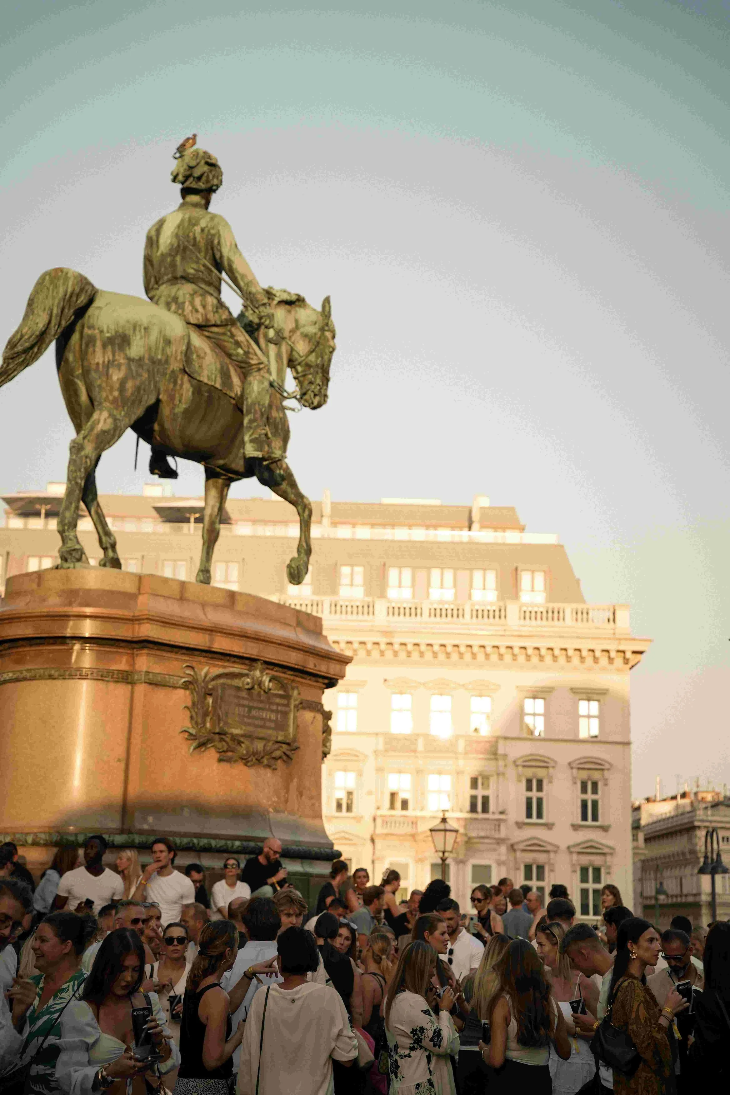 Crowd of people gathered around an equestrian statue in a city square. The statue features a man on horseback. Buildings with multiple windows and rooftops are visible in the background.