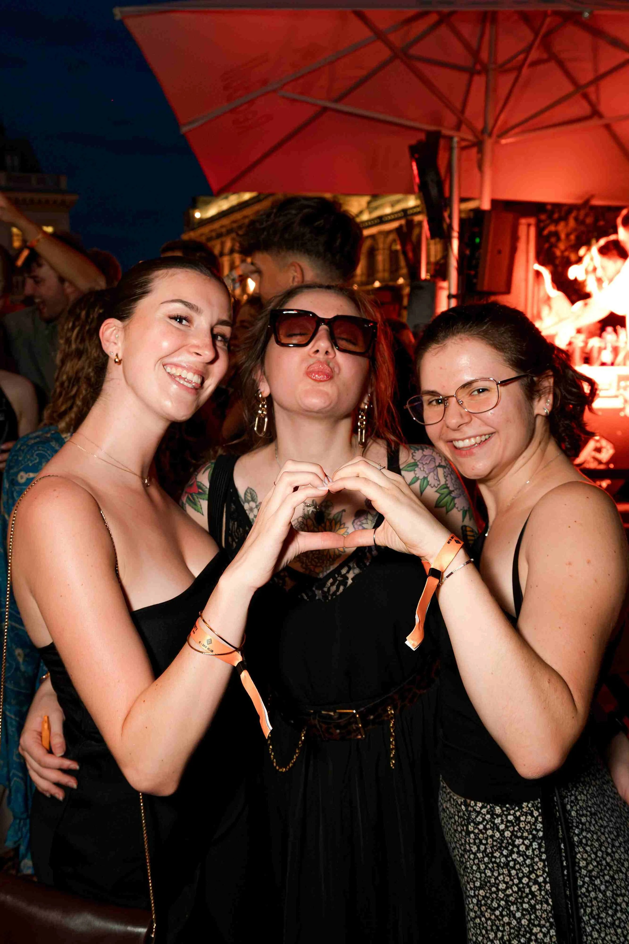 Three women at a night event, holding hands and forming a heart shape with their fingers; smiling and posing for the camera, with a stage and people in the background.