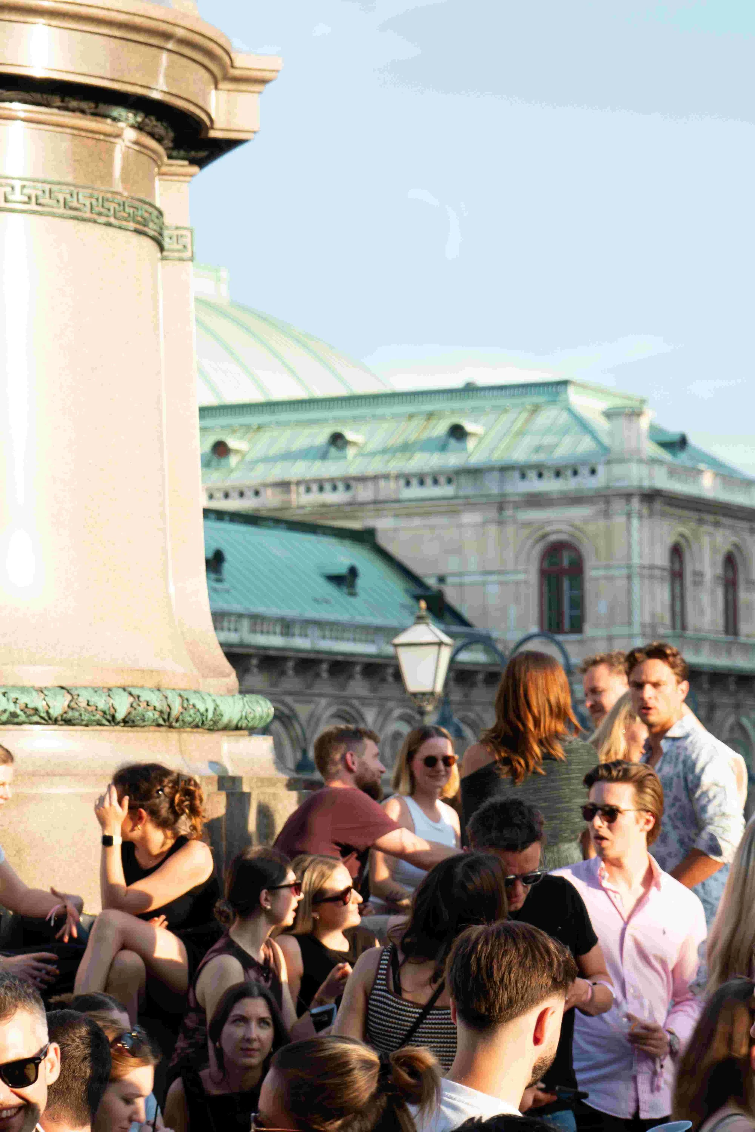 A crowd of people gathered outdoors near a historic building with a green, domed roof and ornate architecture.