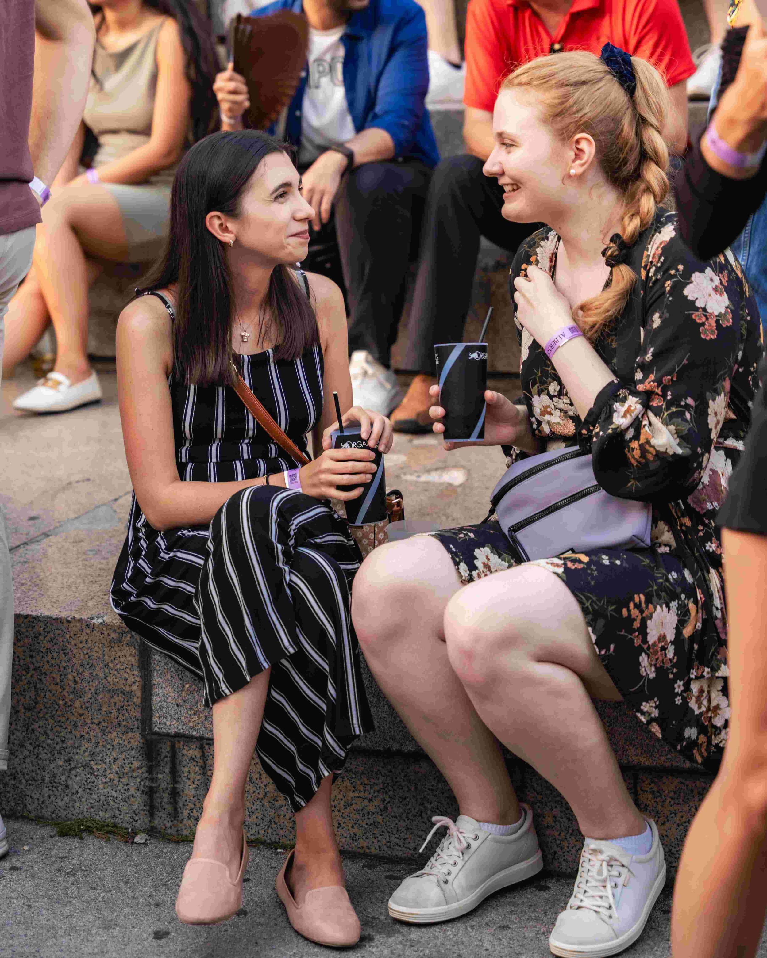 Two young women with light skin sitting on the ground at an outdoor event, smiling at each other. One wears a striped dress and the other wears a floral dress, holding drinks. Several other people are in the background, sitting or standing around.