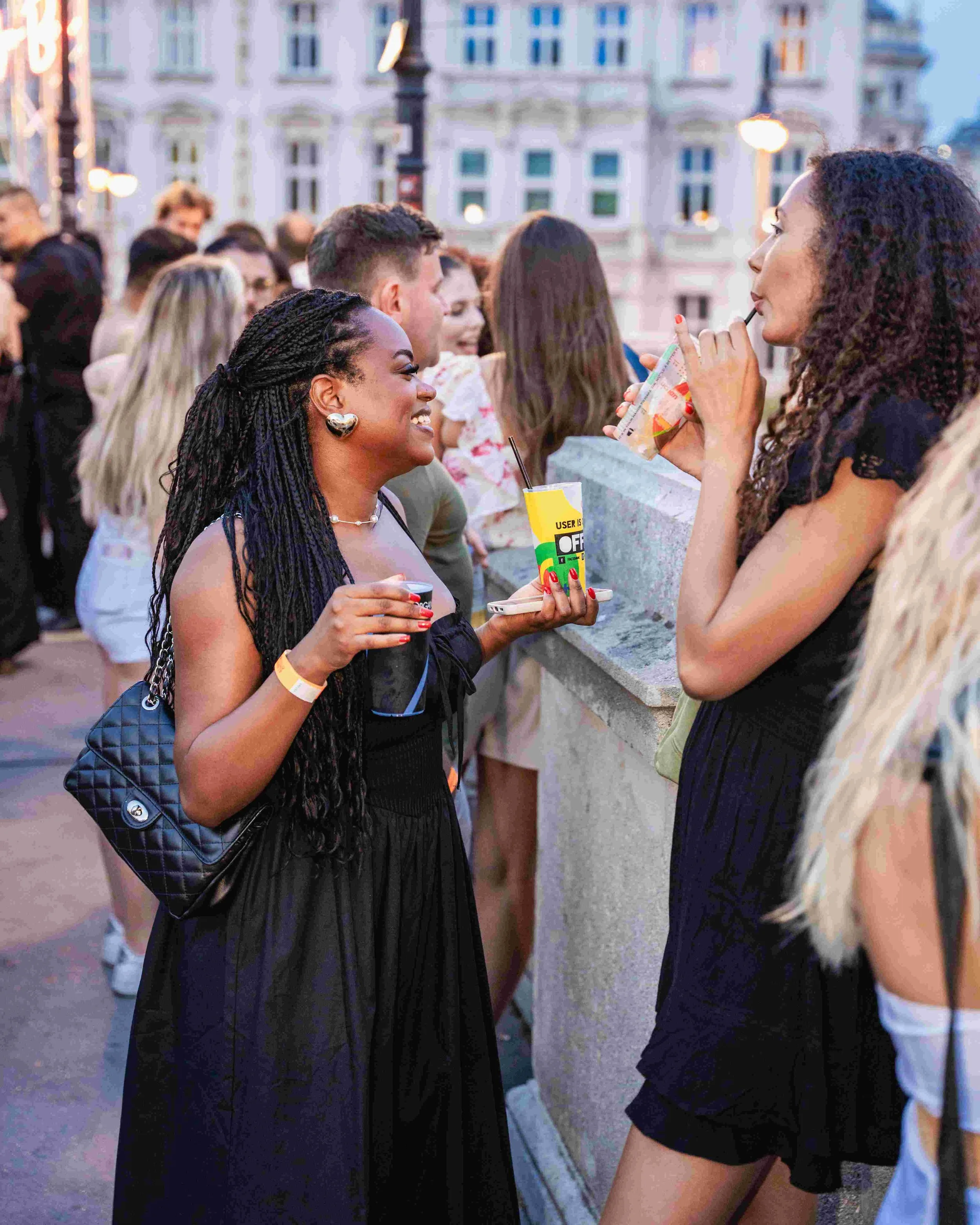 A diverse group of people gathered outdoors in an urban setting during evening, engaged in conversation and enjoying drinks, with classic European architecture in the background.