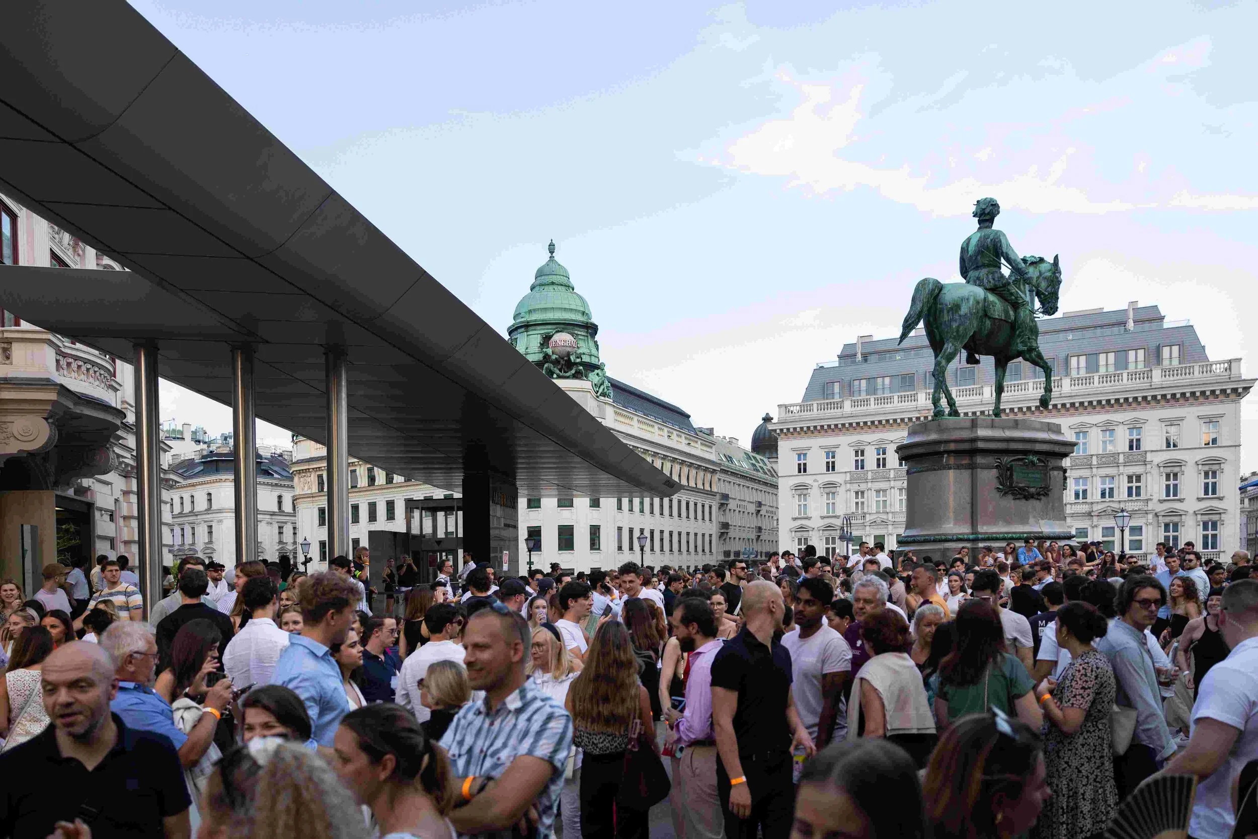 A crowded urban square with many people, with historic buildings and an equestrian statue of a man in the background.