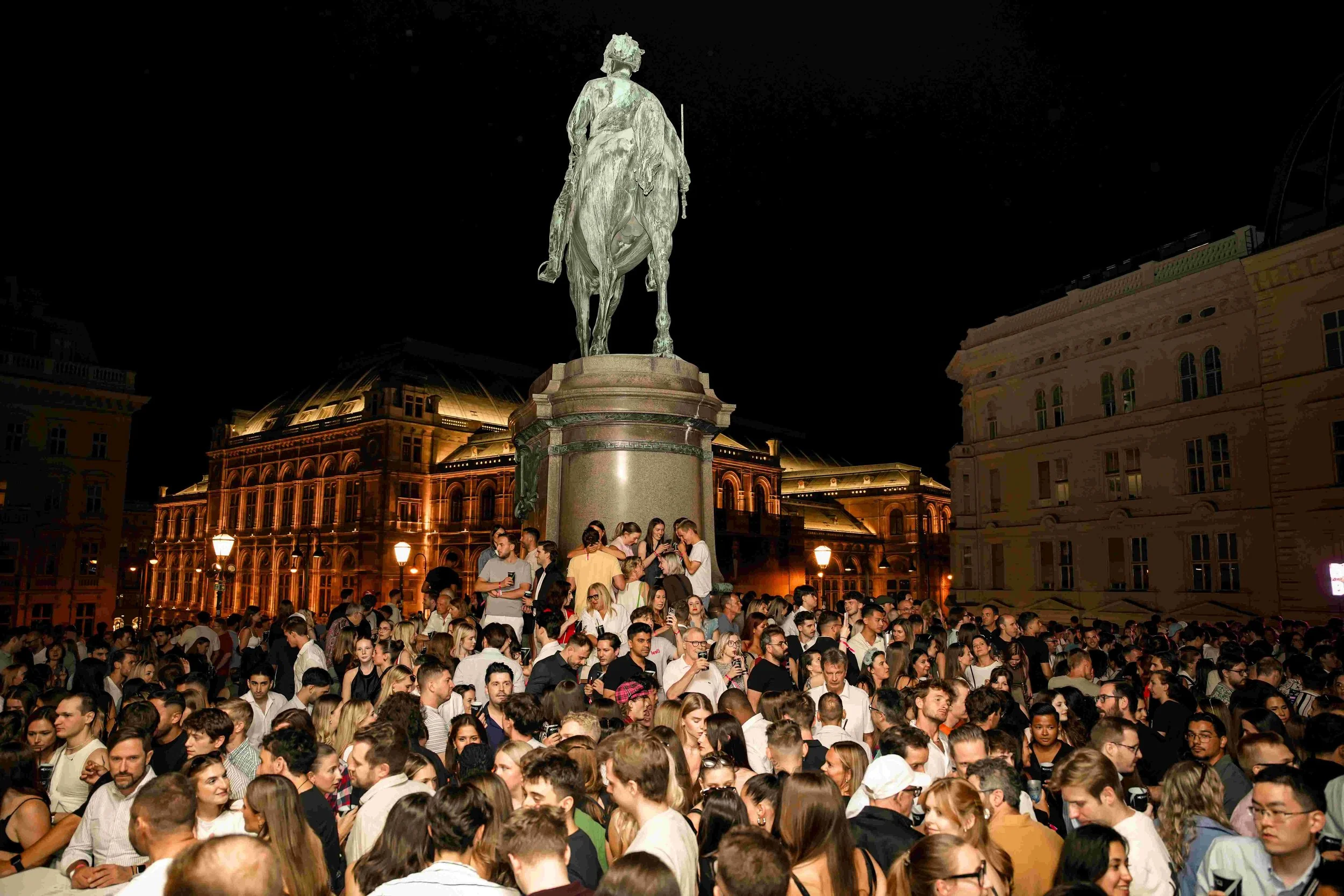 Nighttime scene of a large crowd of people gathered around a statue of a man on horseback.