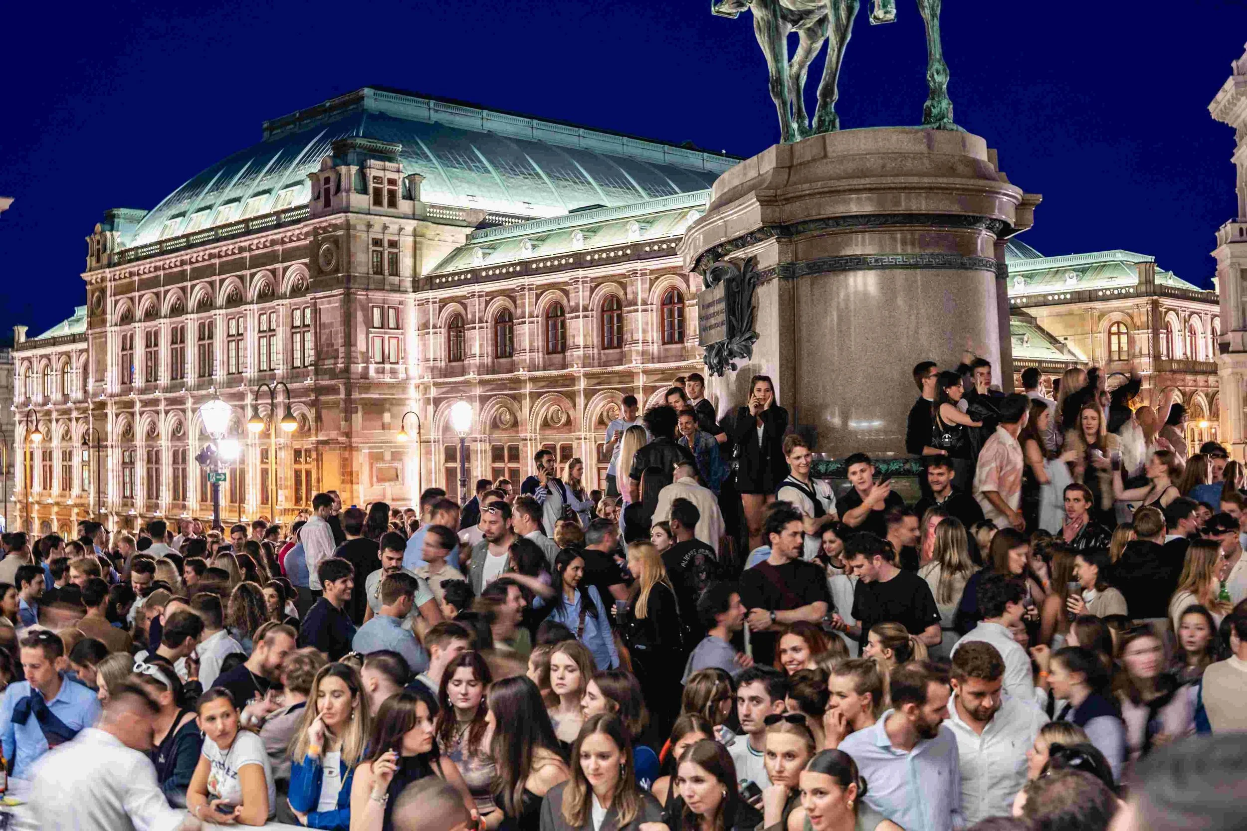 Crowd of people gathered outside at night near historic building with domed roof and art sculpture on a pedestal